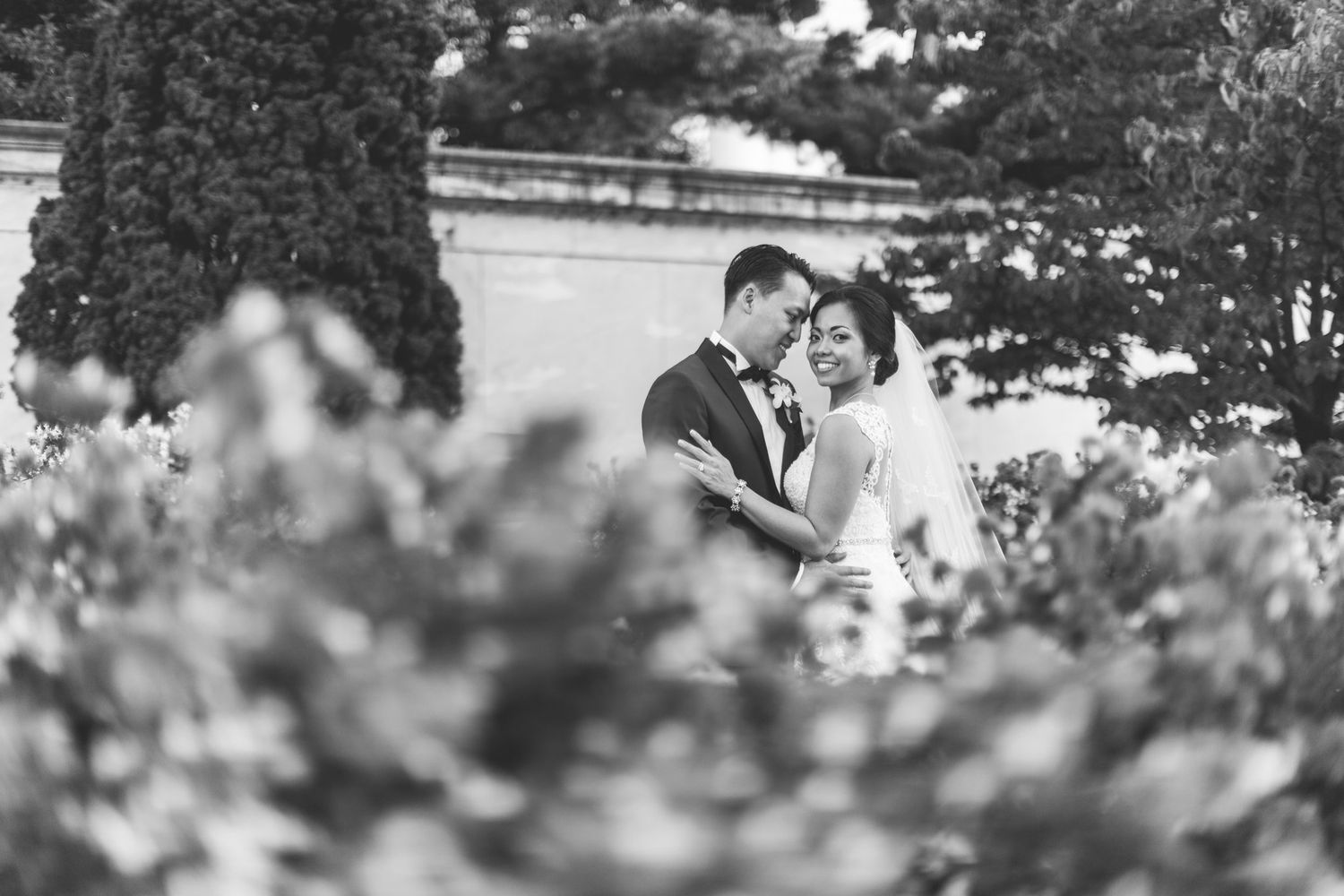 Black and white wedding photo of bride and groom embracing among lush garden greenery near elegant stone architecture.