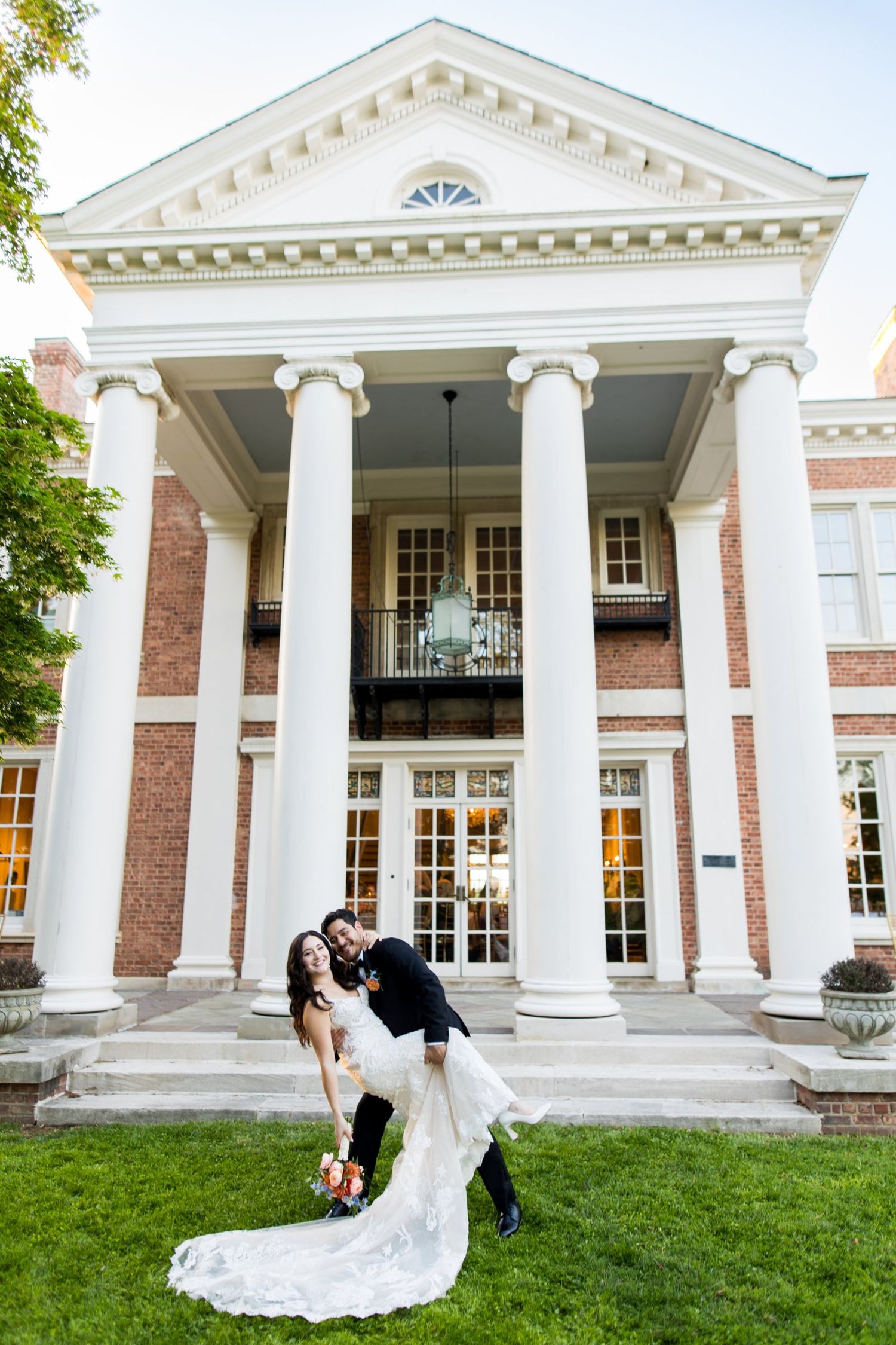 Groom dips bride in white lace gown before a grand brick mansion with white columns on lush green lawn.