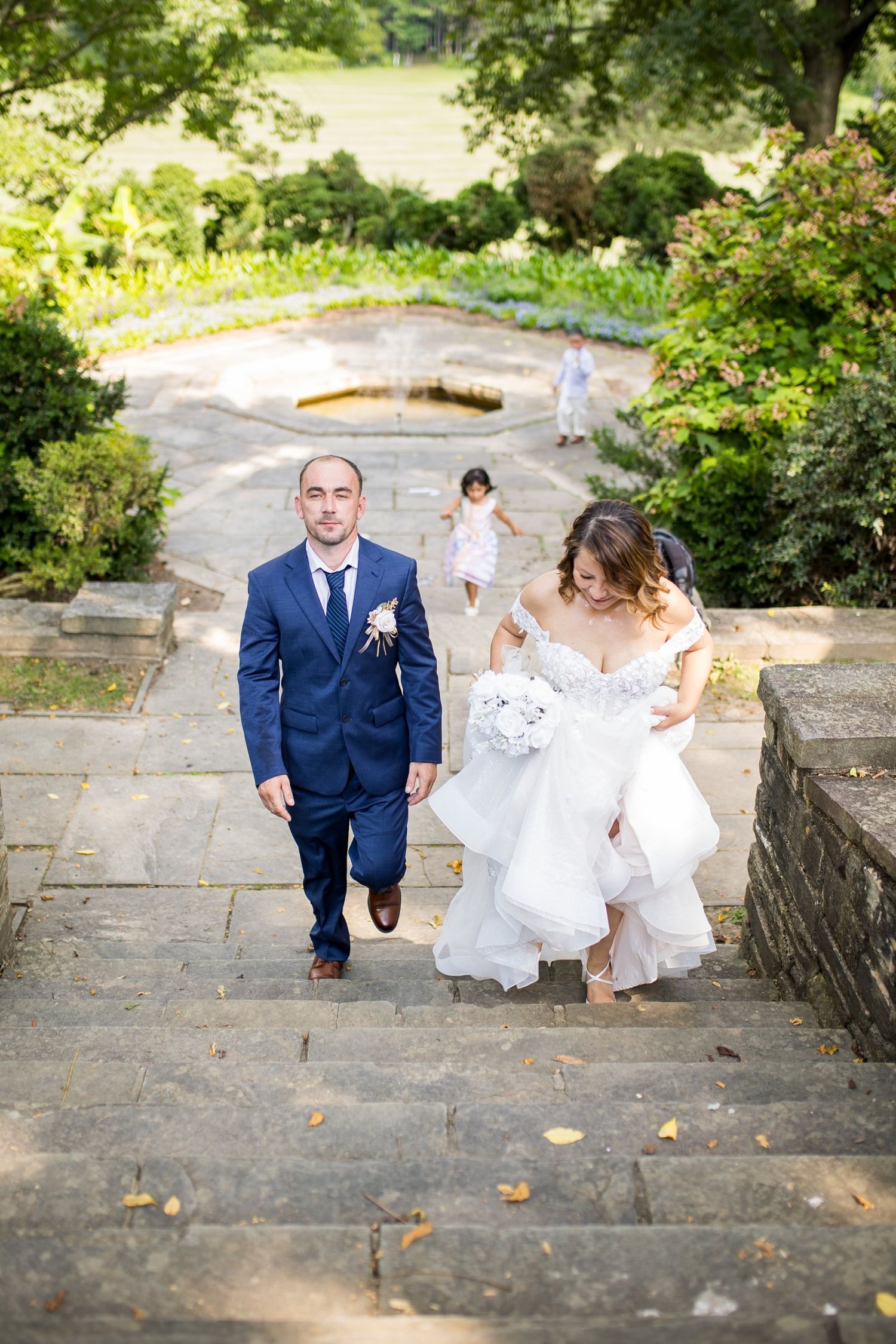 Bride and groom walk up stone steps in lush garden, bride in white gown holding bouquet, groom in navy suit.
