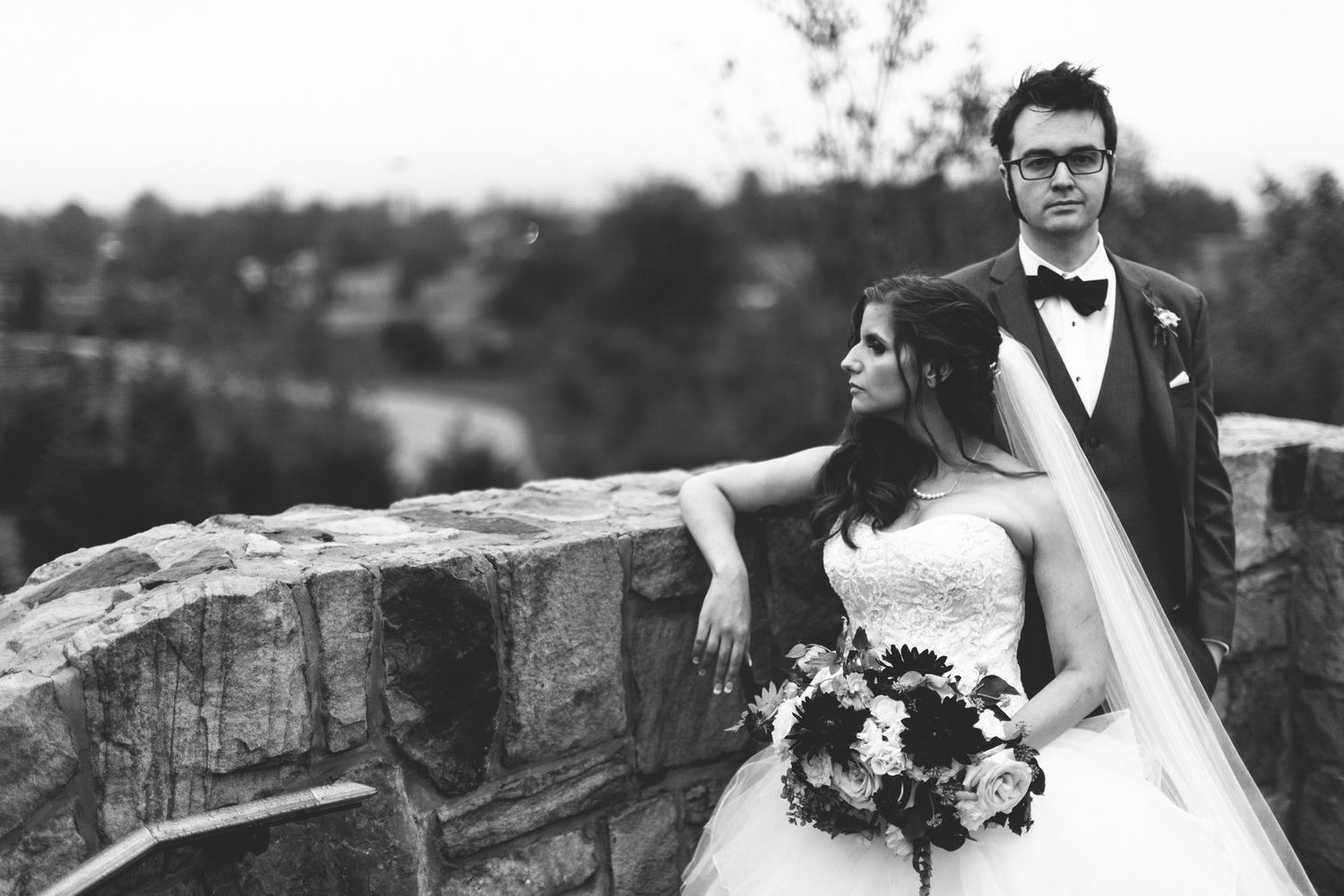 Black and white wedding photo of bride holding bouquet and groom in tuxedo posing on stone wall outdoors.