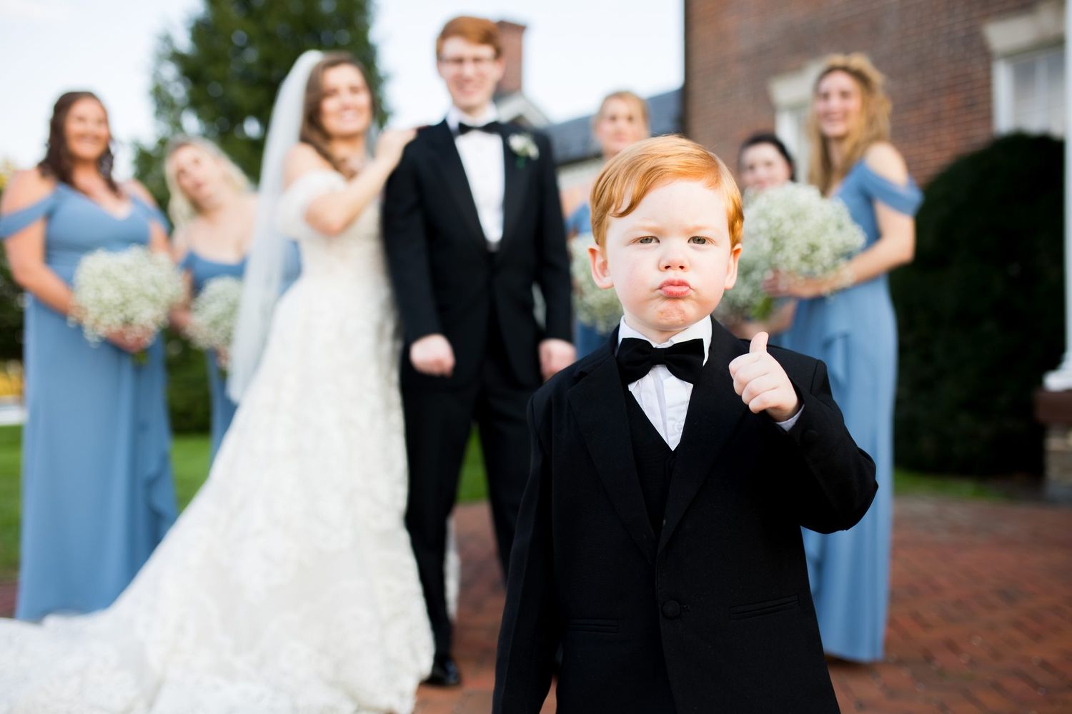 Red-haired ring bearer in tuxedo gives thumbs up in front of wedding party in blue bridesmaid dresses.