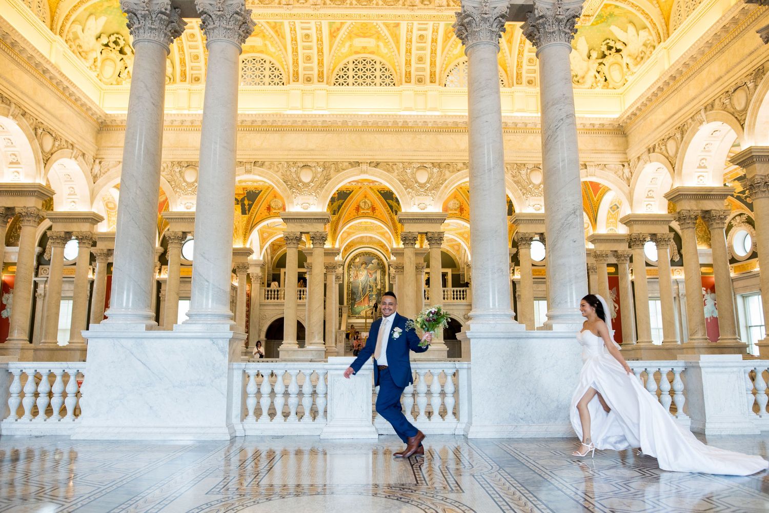 Bride and groom walking through ornate marble columns in a grand wedding venue with golden architecture.