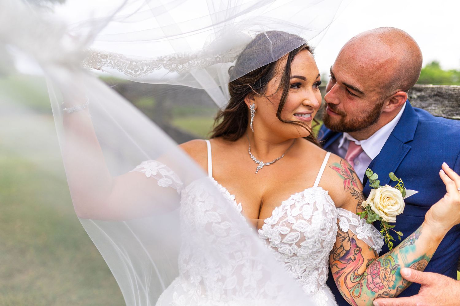 Bride in lace wedding dress with flowing veil embraces tattooed groom in blue suit outdoors.