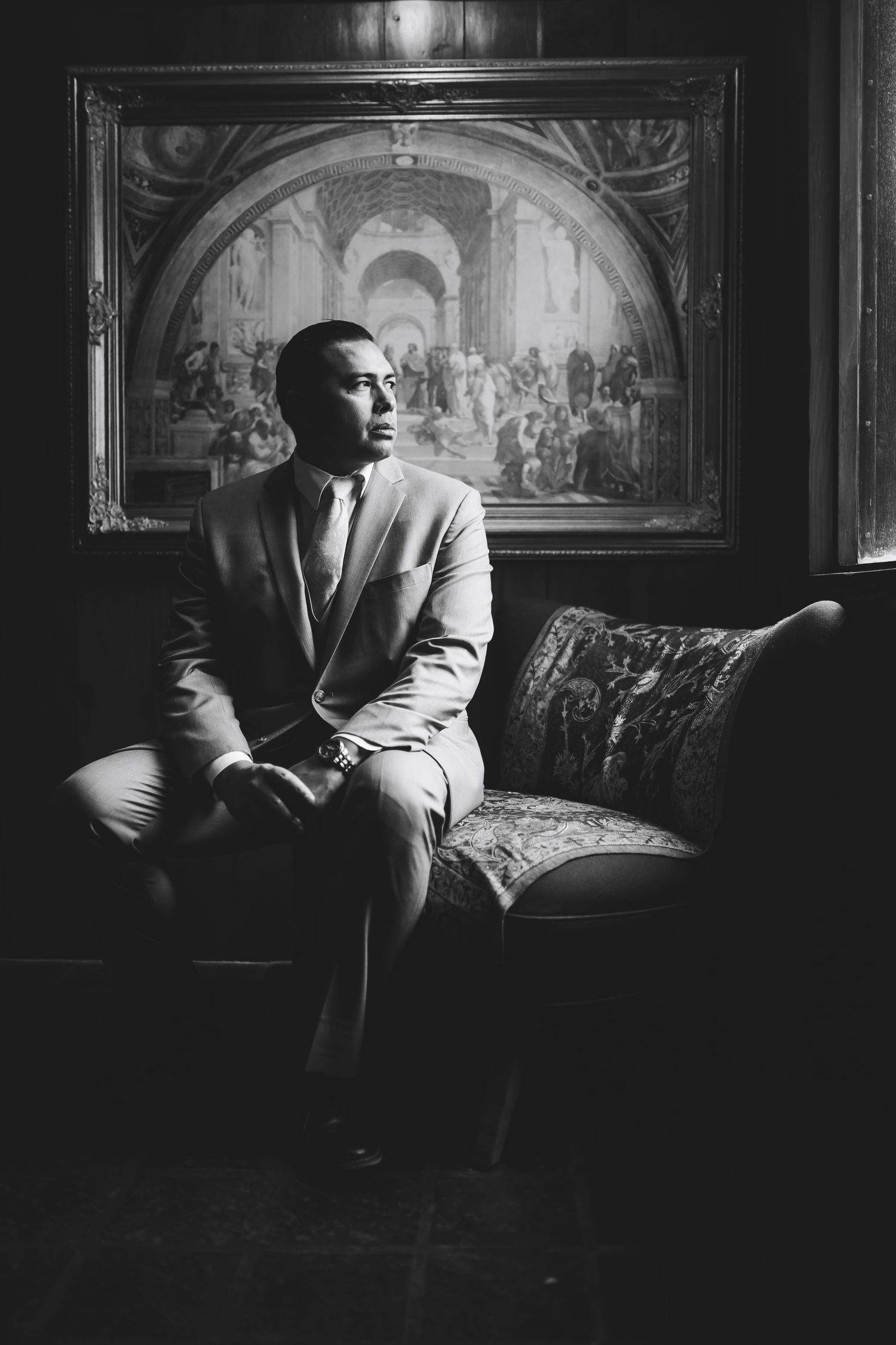 Man in suit sitting on ornate sofa before classic painting in dramatic black and white portrait.