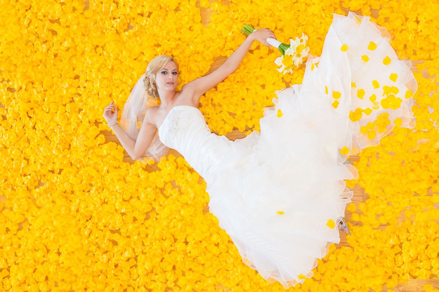 Bride in white gown lying on yellow flower petals, holding bouquet overhead, aerial view wedding photo.