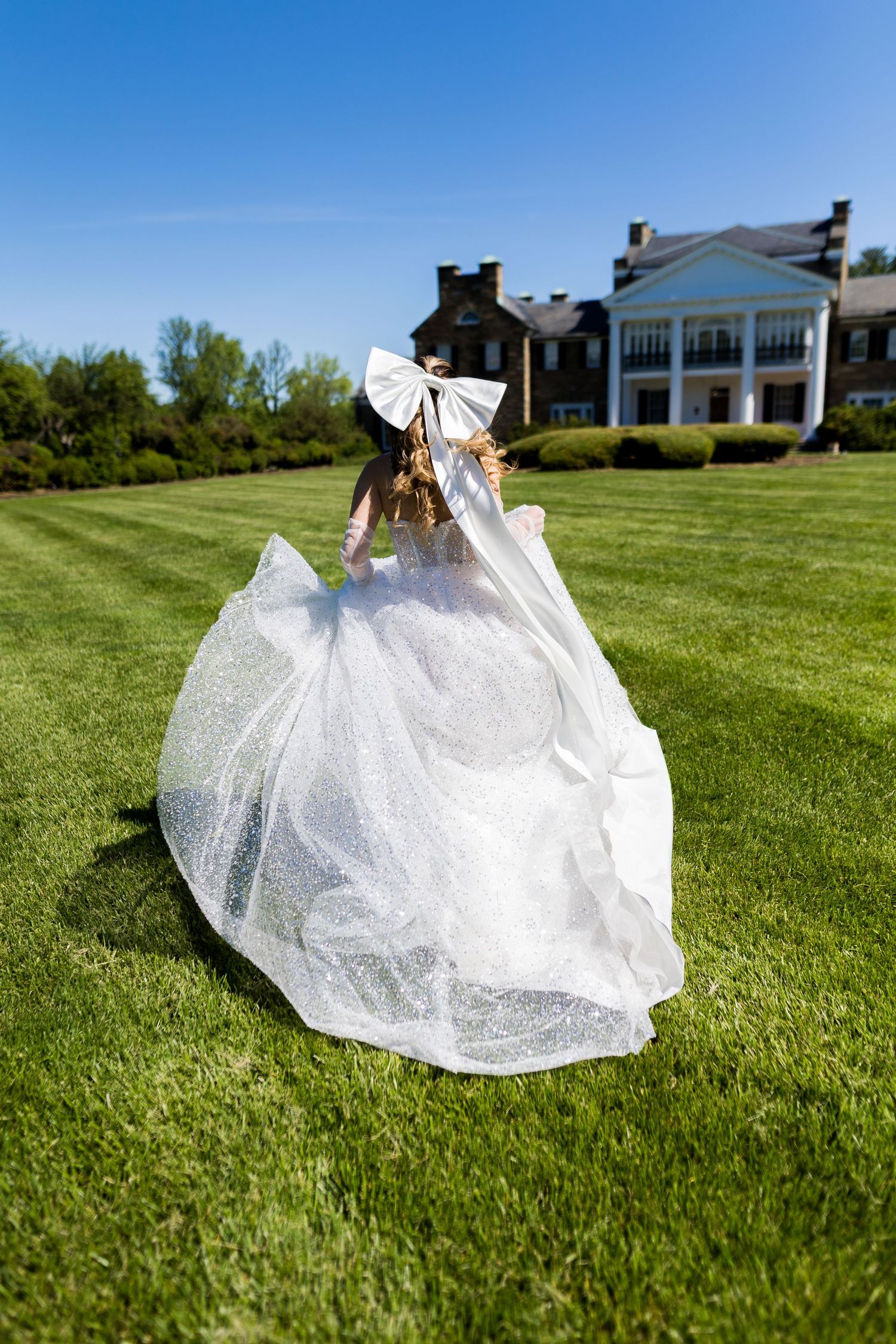 Bride in flowing white gown and bow veil runs across lush green lawn toward elegant brick mansion on sunny day.