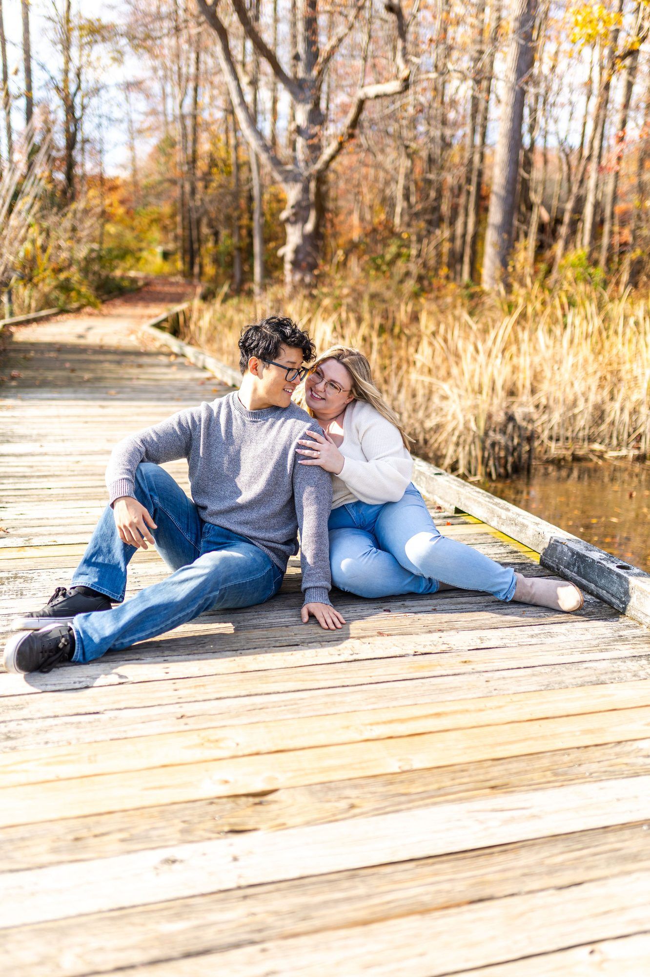 Couple sitting together on a wooden dock surrounded by autumn foliage, sharing a romantic moment by a serene pond.