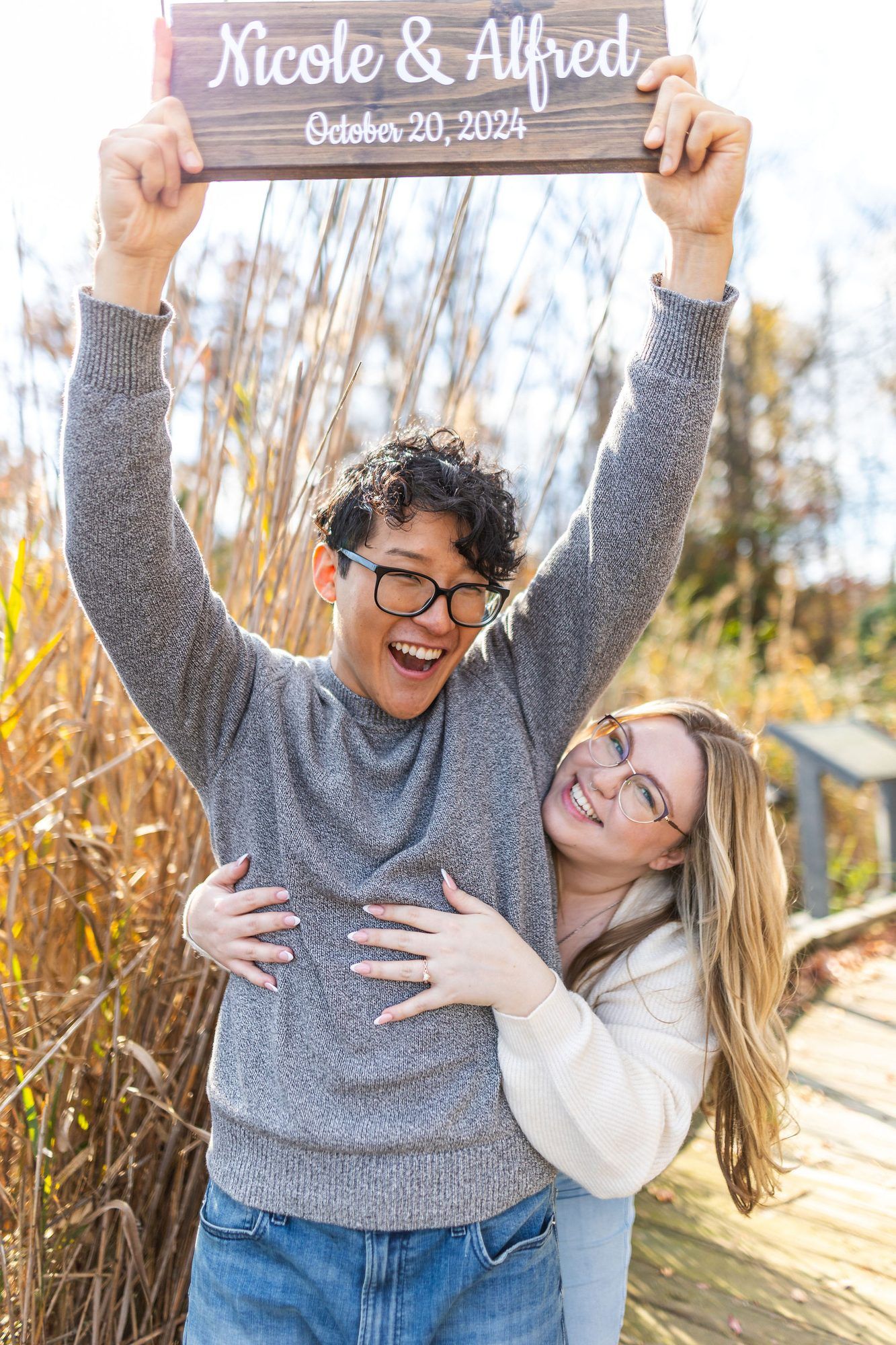 Happy couple holding a wooden wedding sign reading Nicole & Alfred, October 20, 2024, outdoors in autumn.