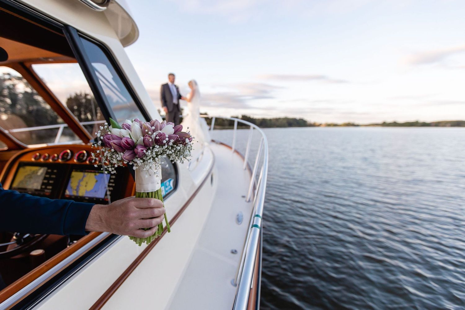Wedding couple stands on luxury yacht deck at sunset while guest holds bridal bouquet over water.