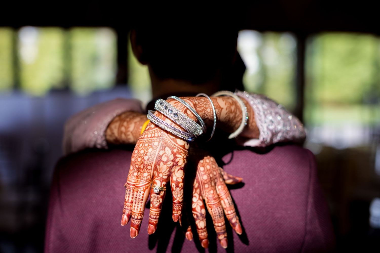 Bride's hands adorned with intricate henna mehndi designs and gold bangles resting on groom's shoulder.