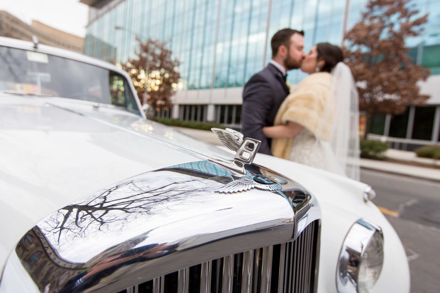 Bride and groom kissing behind a classic white Rolls-Royce at their wedding venue.
