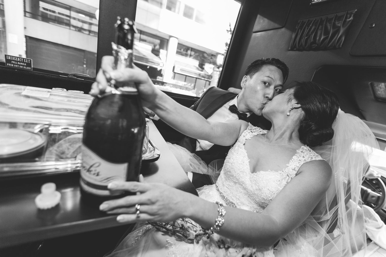 Black and white photo of a bride and groom kissing while holding a champagne bottle in a vehicle on their wedding day.