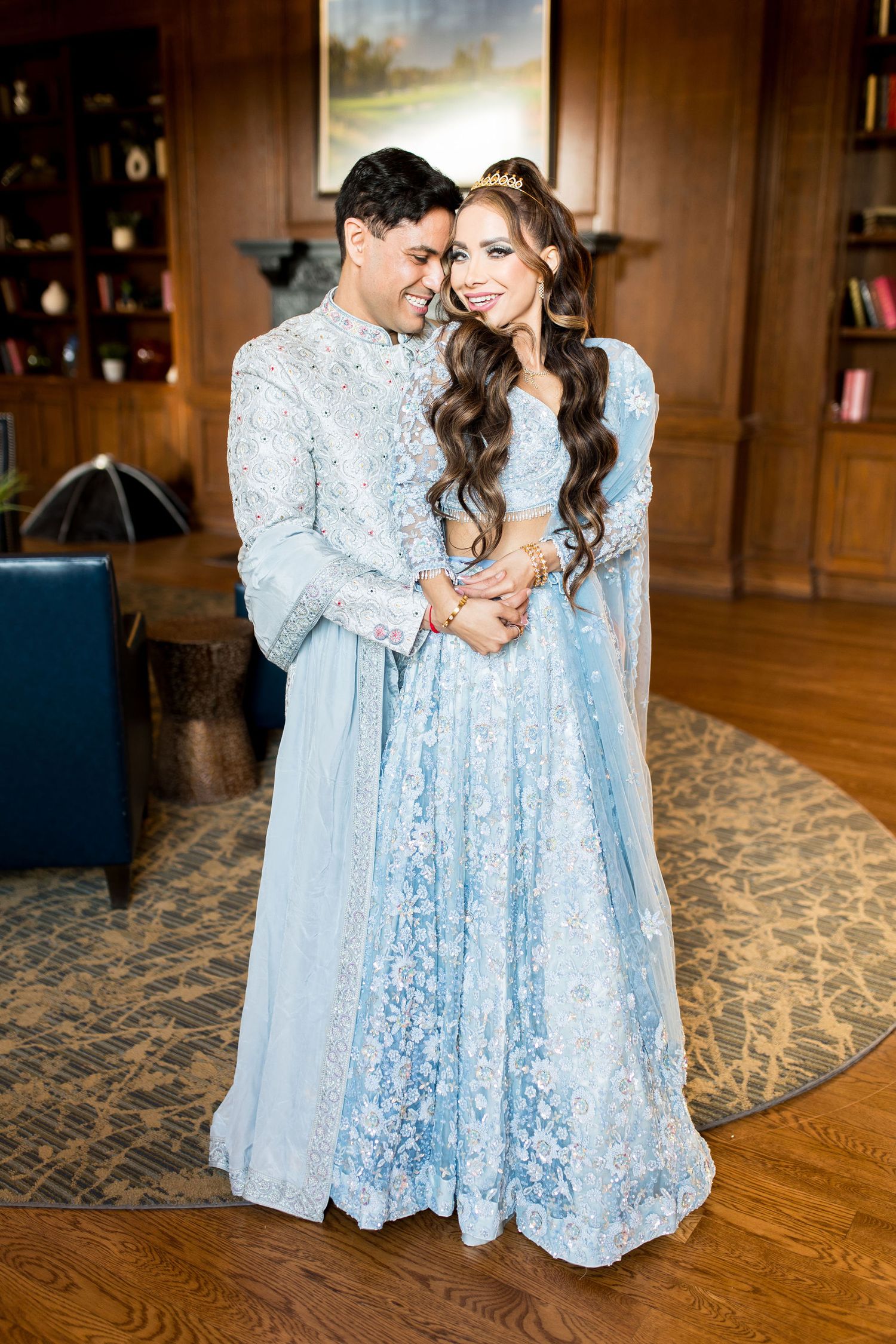 Couple in matching light blue embroidered Indian wedding attire smiling together in an elegant wood-paneled room.