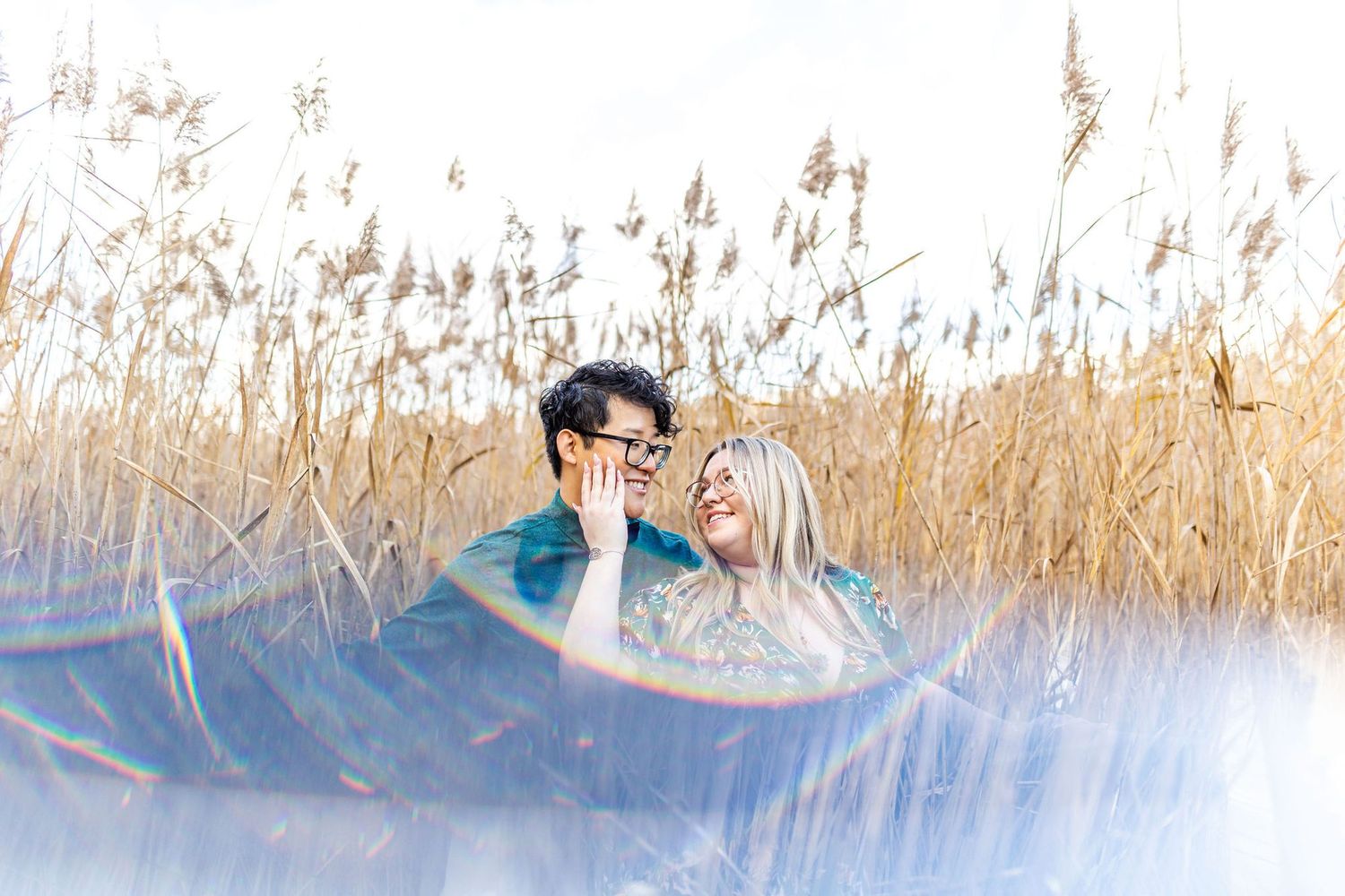 Couple laughing together among golden reeds, captured through a dreamy lens flare effect outdoors.