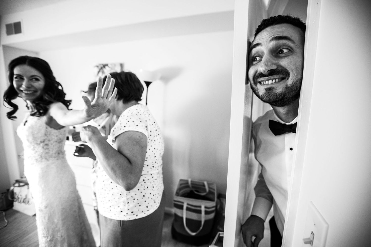 Groom peeks through door smiling as bride gets ready with help in black and white wedding photo.