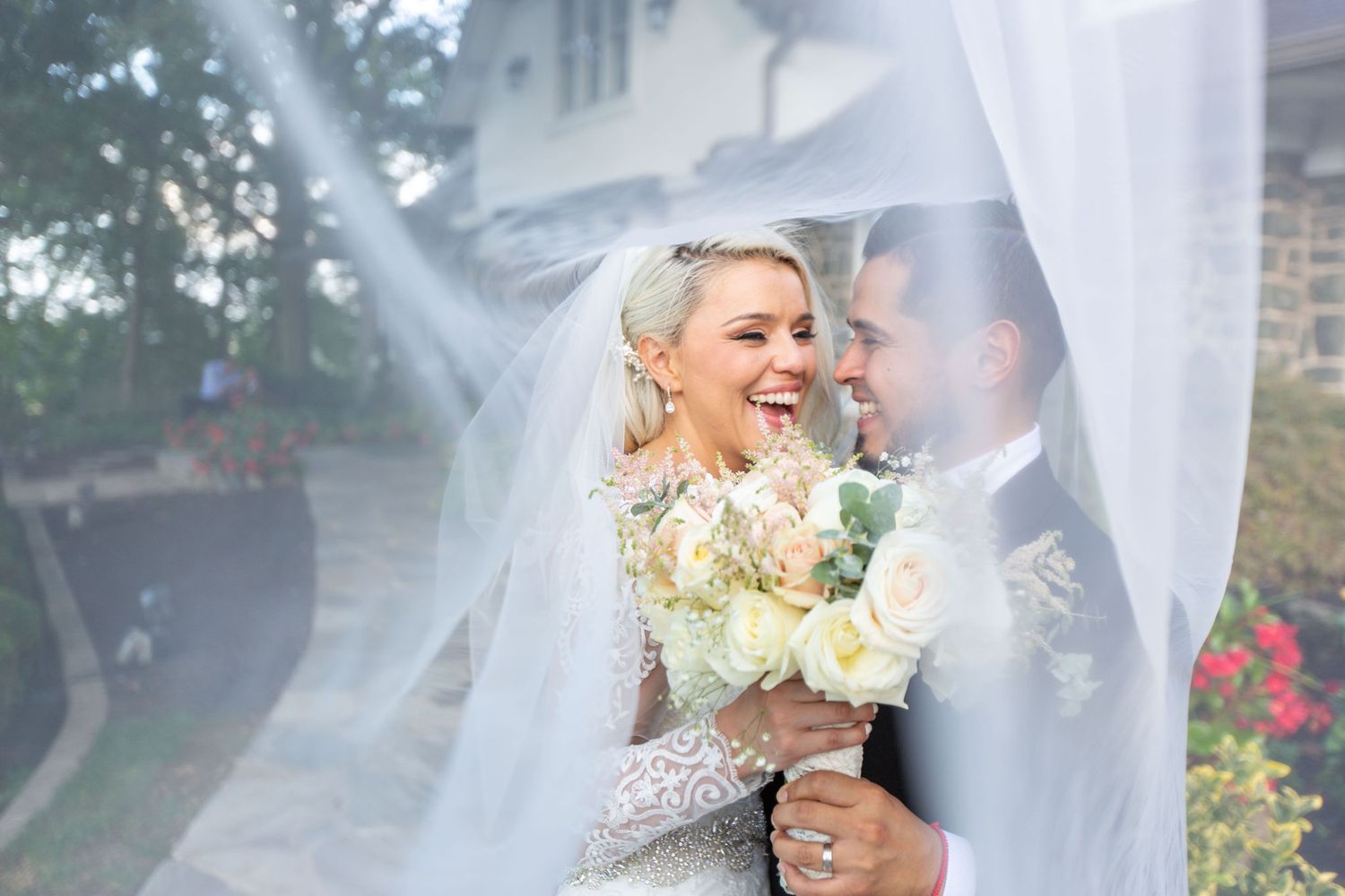 Bride and groom laughing under a flowing white veil, bride holding white bouquet in elegant lace wedding dress.