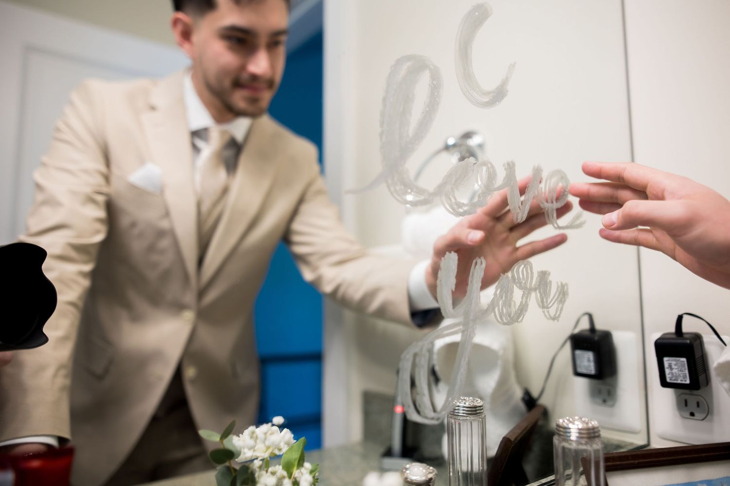 Groom in beige suit placing a wedding ring on bride's hand near a mirror with love decor.