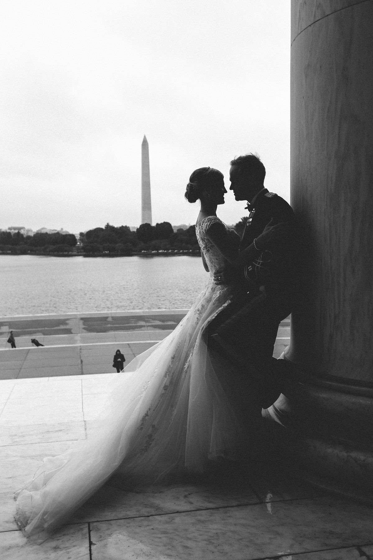 Black and white photo of bride and groom embracing near a column with Washington Monument in background.