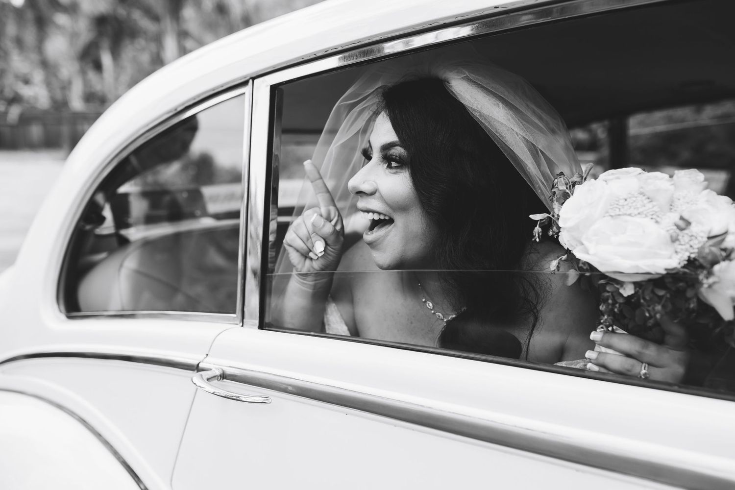 Black and white photo of a smiling bride holding a bouquet, leaning out of a vintage white car window on her wedding day.