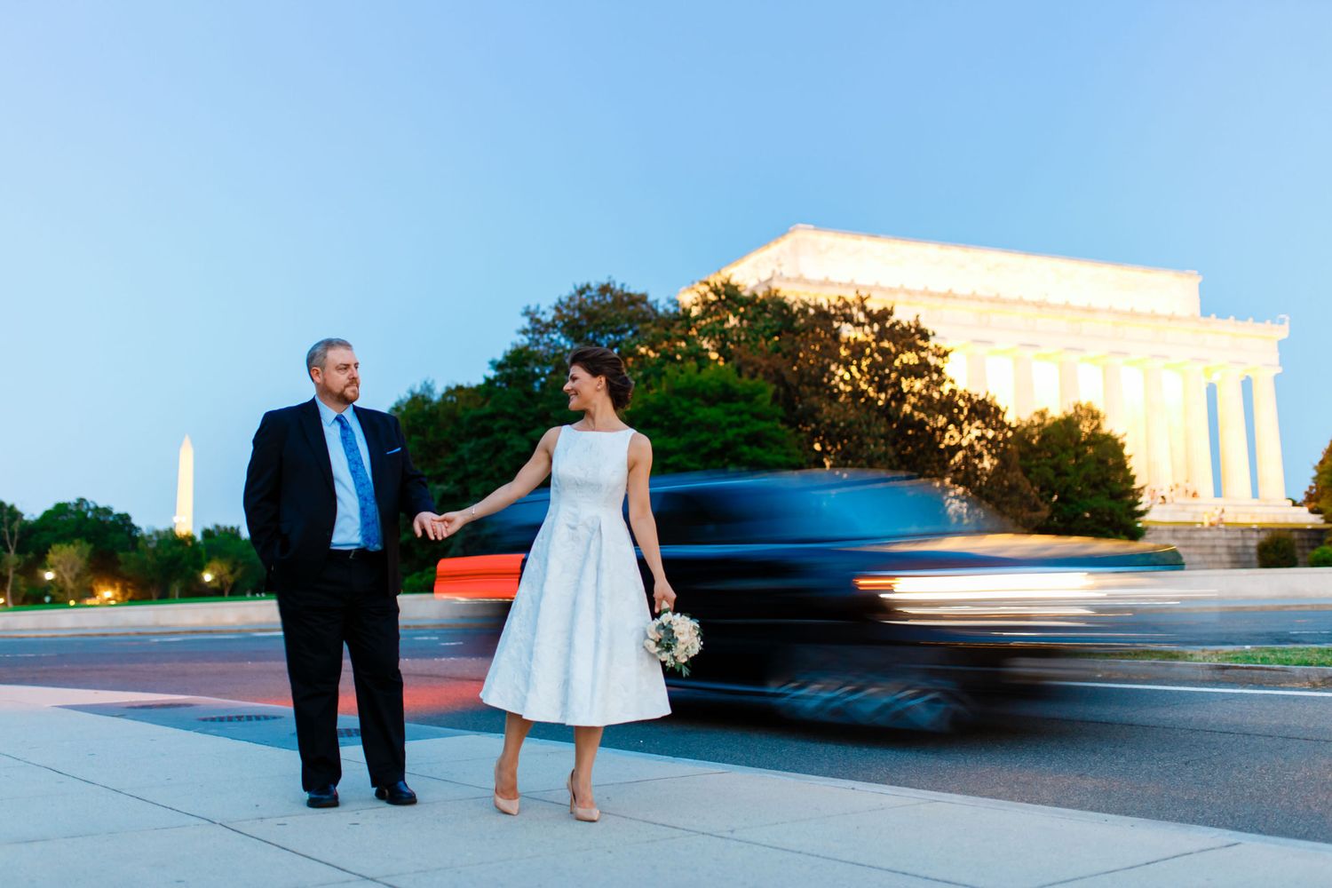 Bride and groom hold hands near the Lincoln Memorial and Washington Monument at dusk in Washington DC.