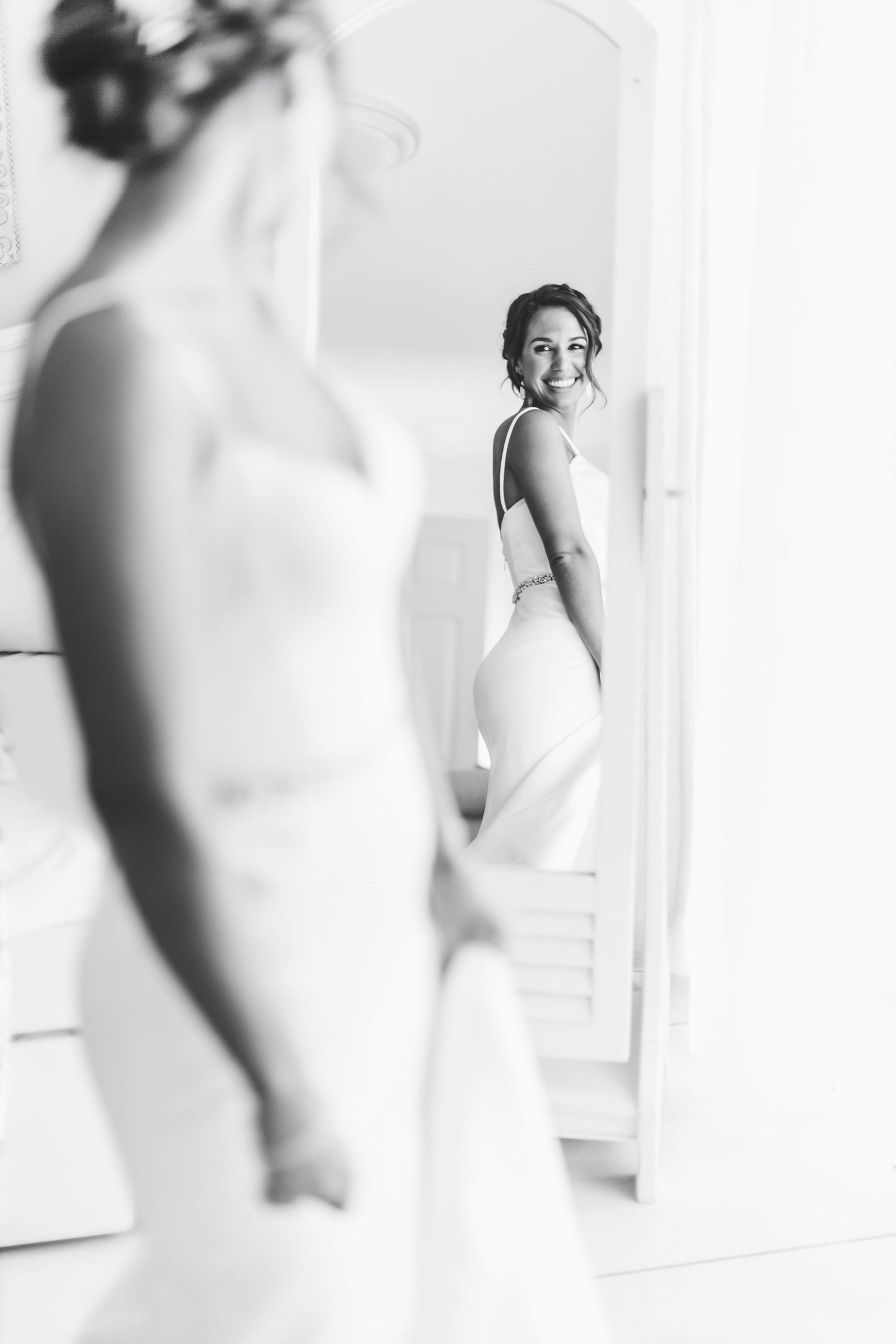 Black and white photo of a smiling bride in a white gown admiring her reflection in a full-length mirror.