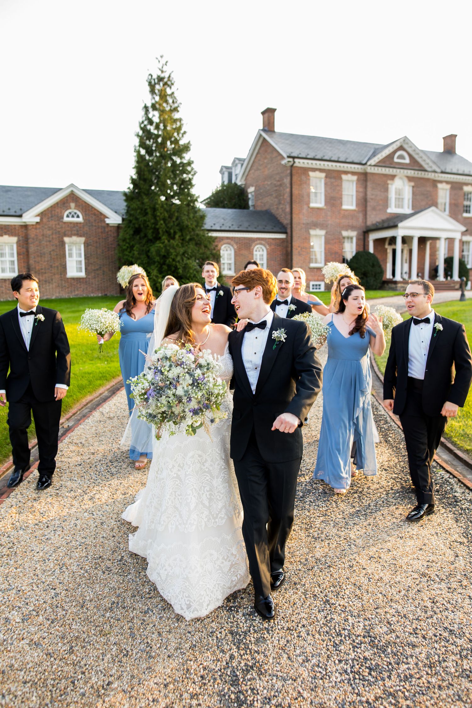 Bride and groom walk with wedding party in blue gowns and black tuxedos outside a Belmont Country Club.