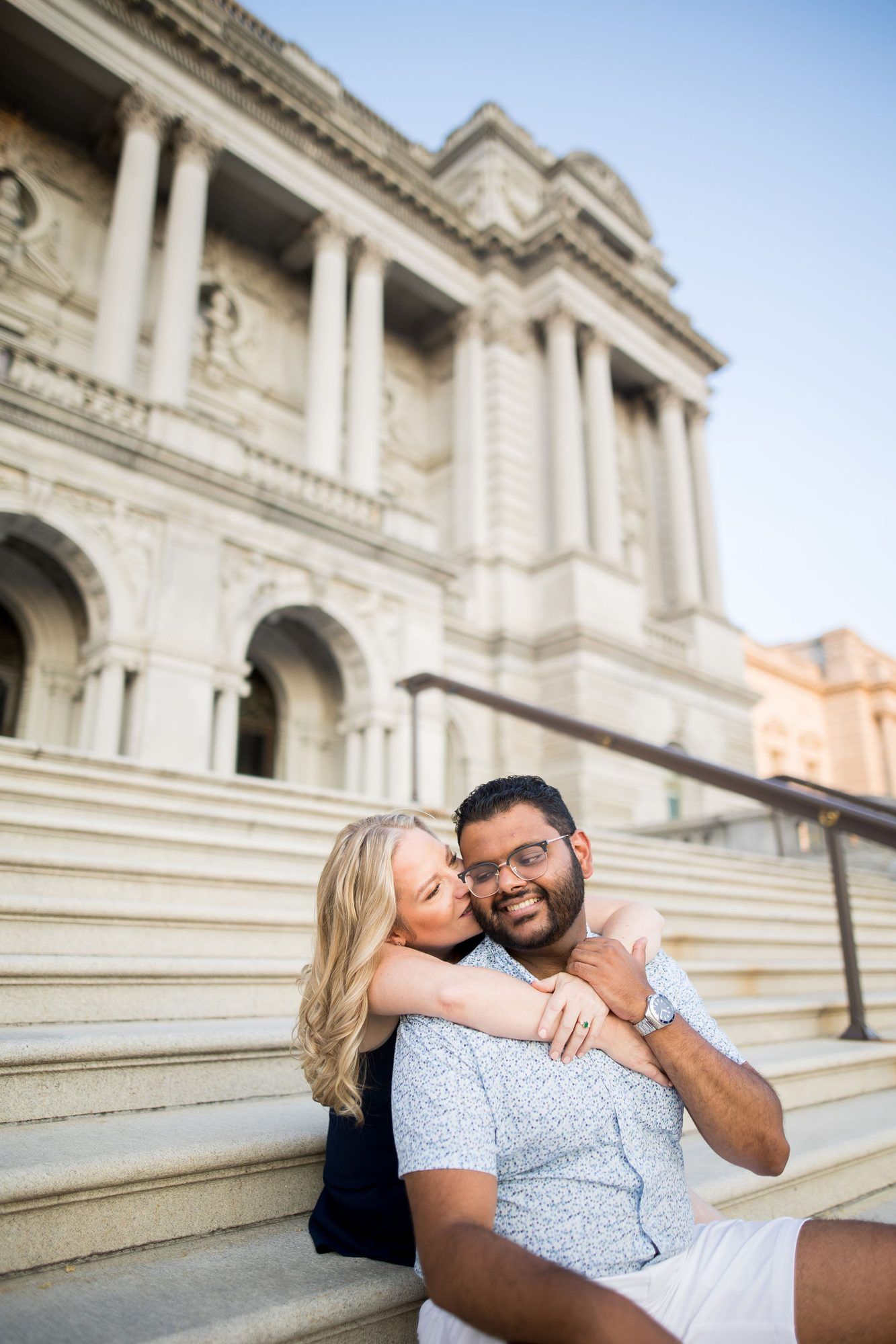 Couple sitting on steps of a grand classical building, woman kissing man on cheek while embracing him.