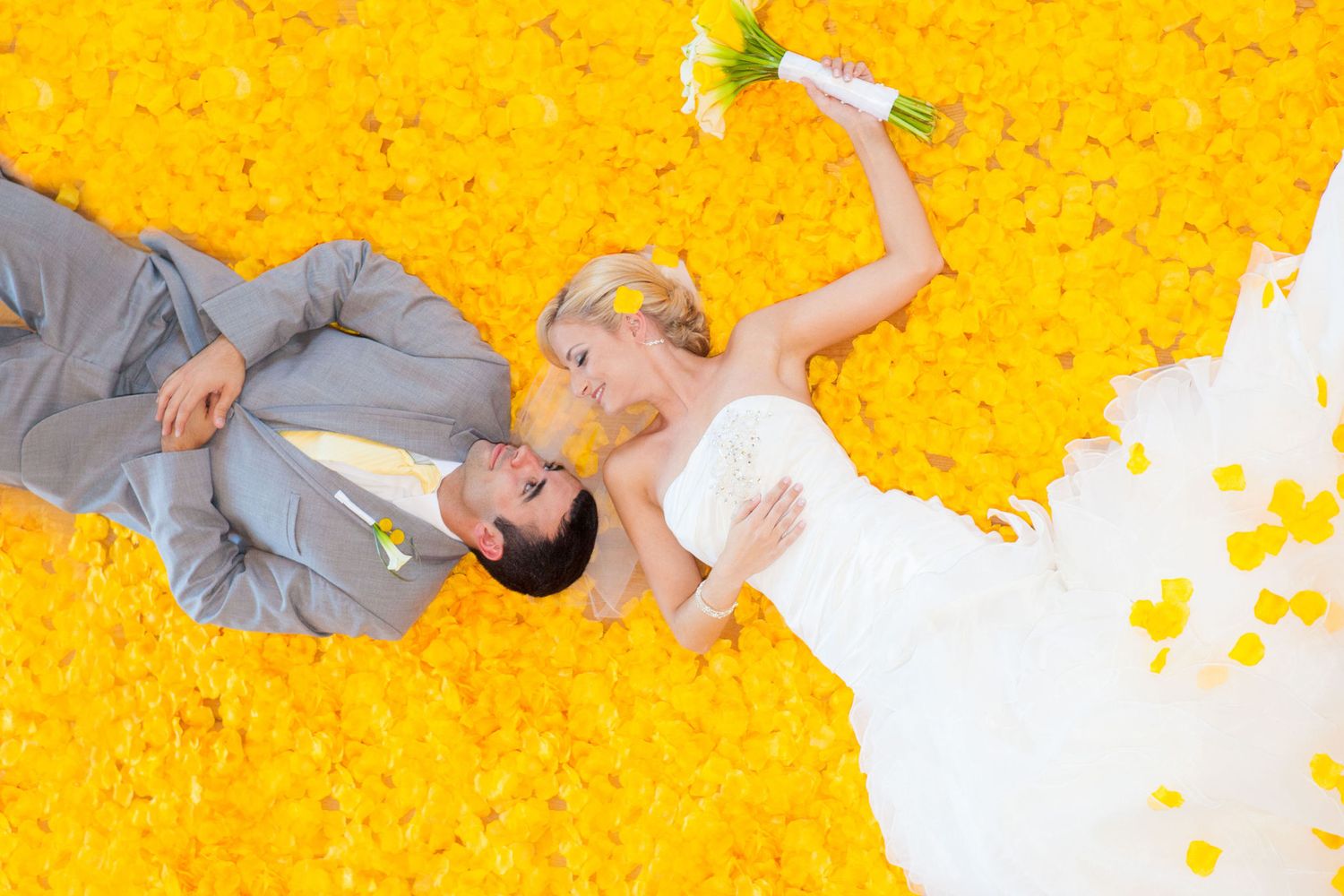 Bride and groom lying on vibrant yellow flower petals, bride holding bouquet aloft in stunning wedding photo.