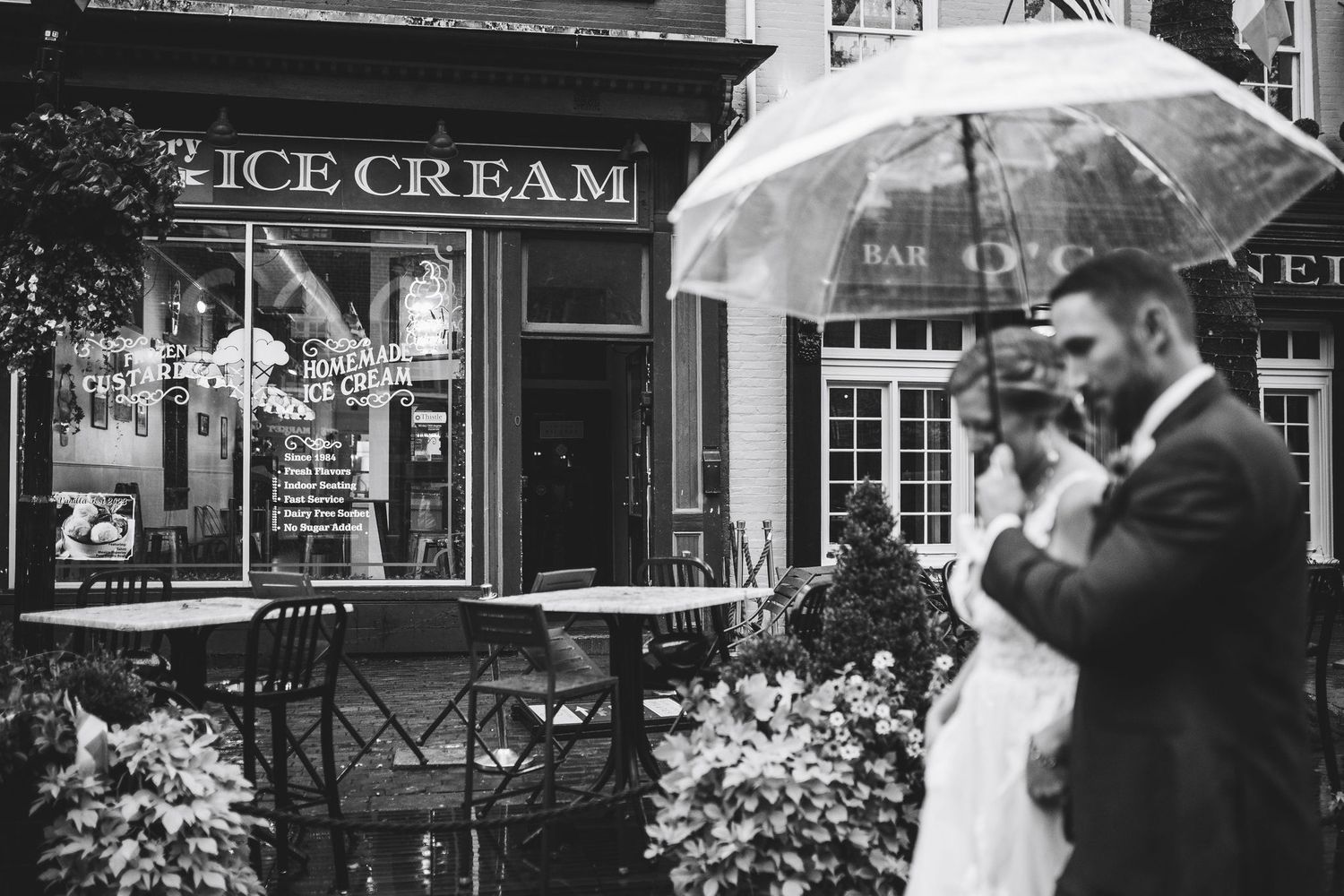 Black and white photo of a bride and groom sharing an umbrella in the rain outside an ice cream shop.