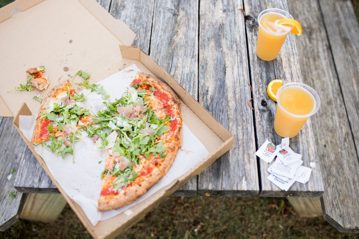 Half-eaten pizza in a cardboard box with arugula toppings on a wooden picnic table beside two orange juice drinks.