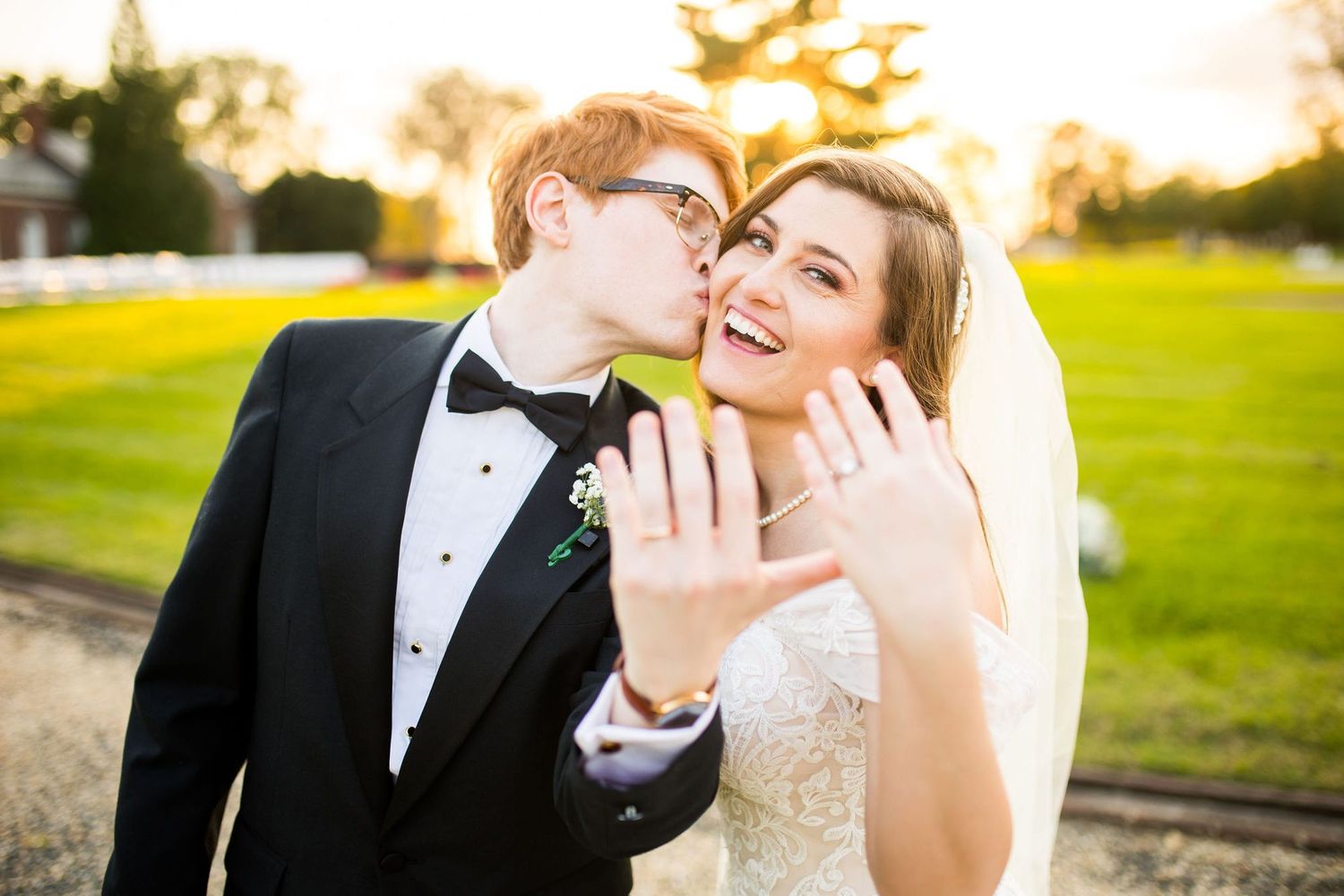 Newlywed couple showing wedding rings, groom kissing bride's cheek in tuxedo and white gown outdoors.