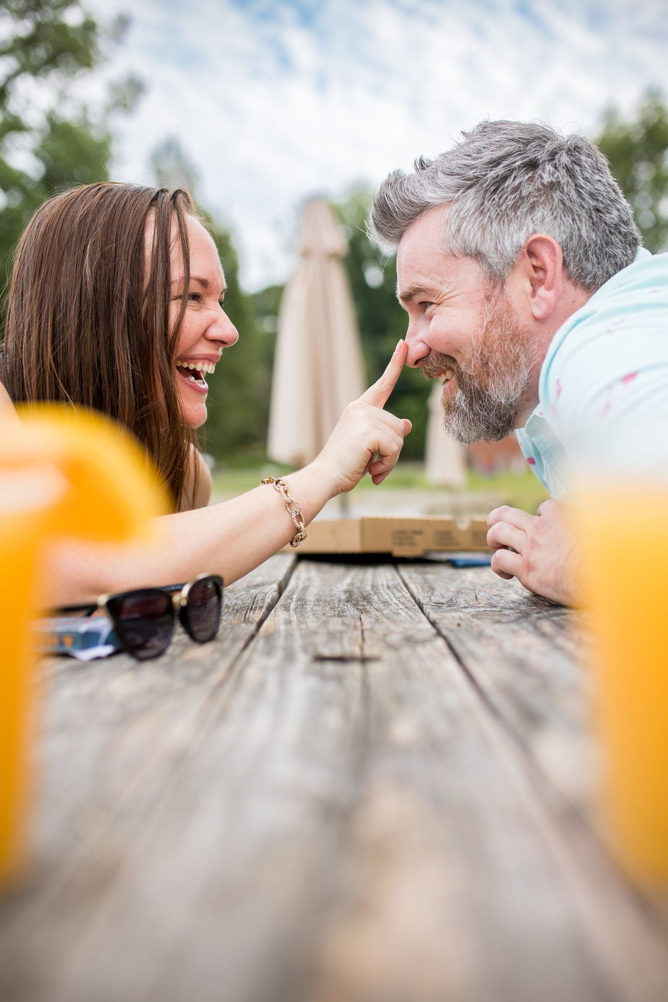 Couple laughing at outdoor picnic table, woman playfully touching man's nose, with orange drinks in foreground.