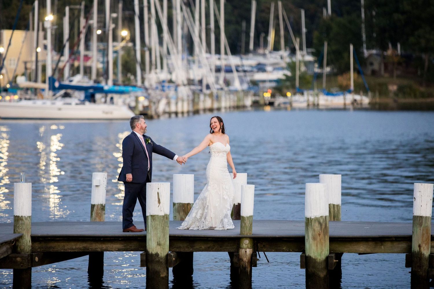 Bride and groom hold hands on a wooden dock at a marina with sailboats at sunset on their wedding day.