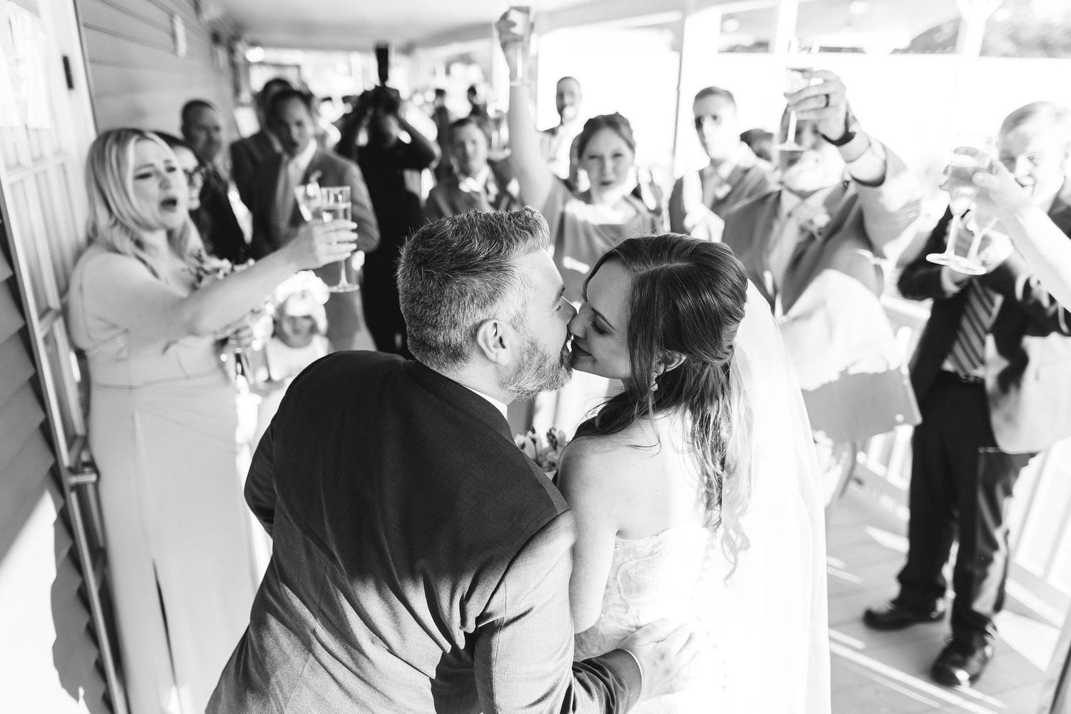 Black and white photo of a couple sharing a kiss at their wedding reception surrounded by cheering guests raising glasses.