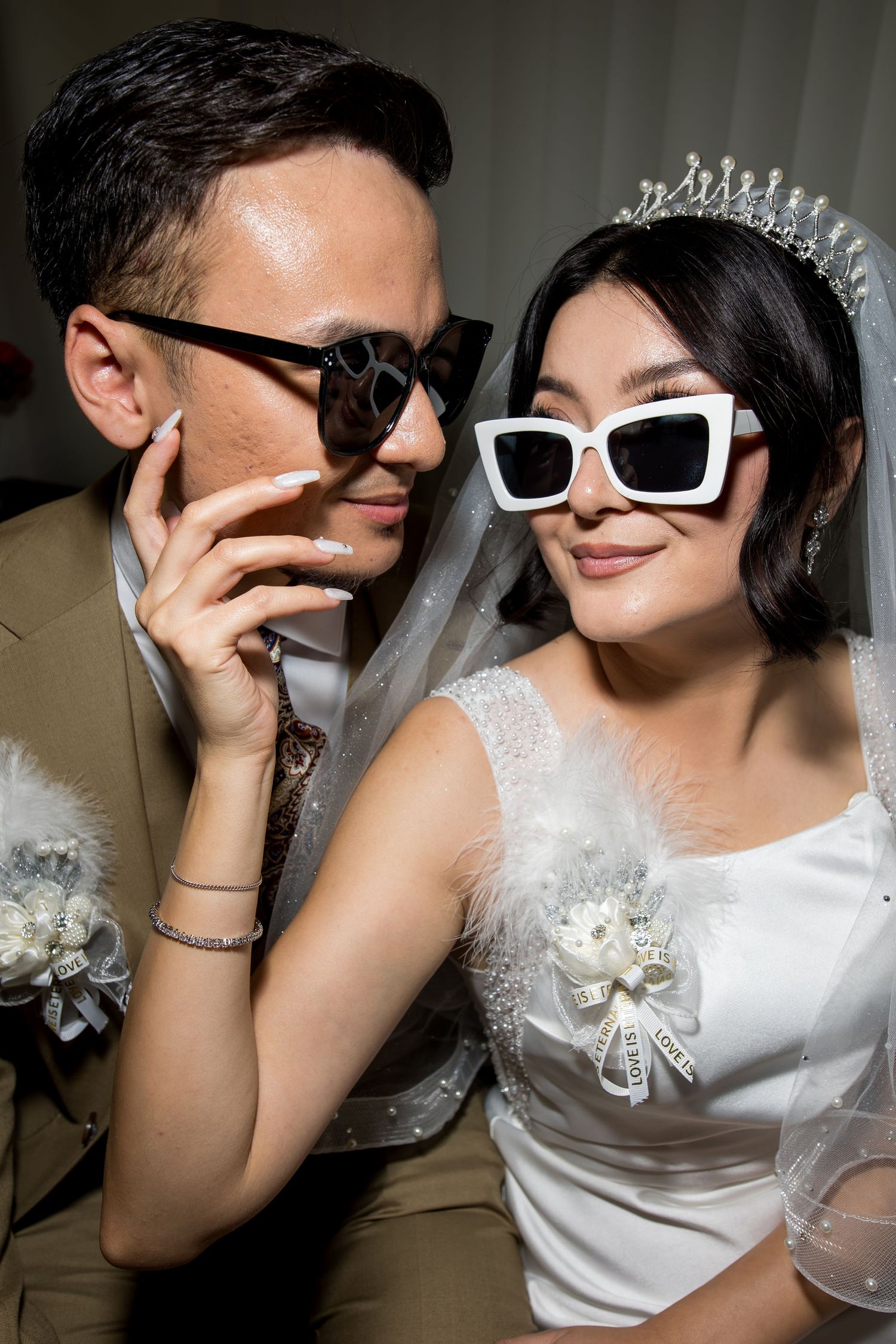 Stylish newlywed couple posing in sunglasses, bride wearing white dress, tiara, and veil on wedding day.