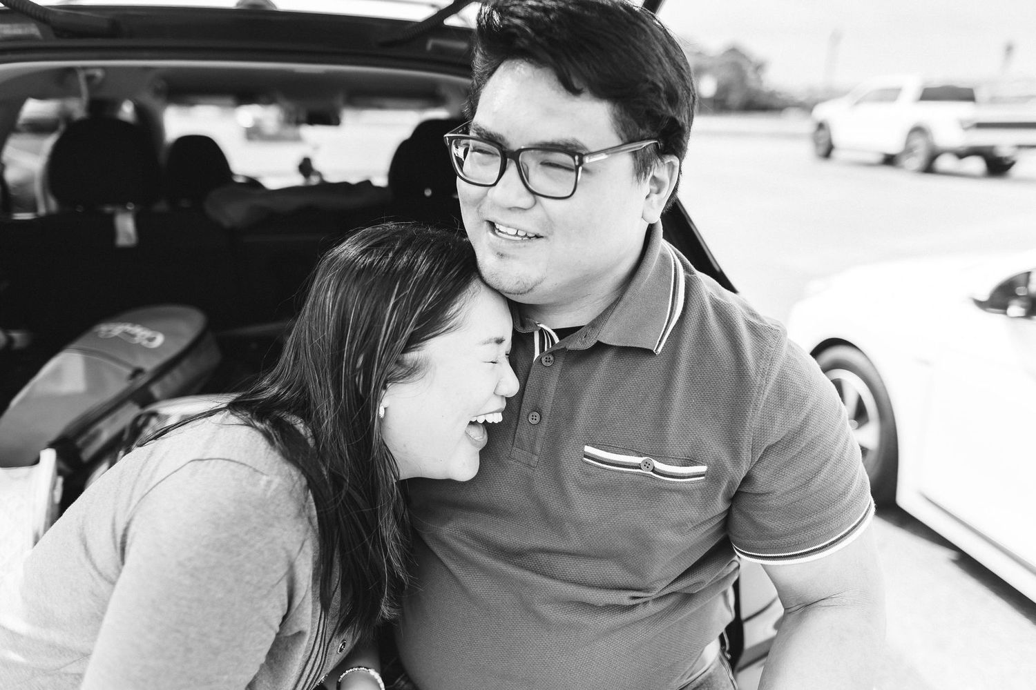 Black and white photo of a smiling couple leaning together near a car trunk in a parking lot.