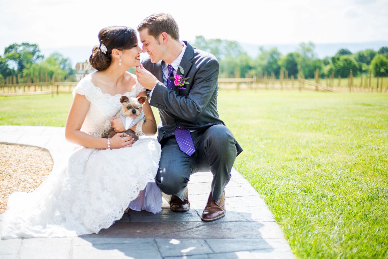 Bride and groom crouching on stone path holding small dog, smiling at each other on their wedding day outdoors.