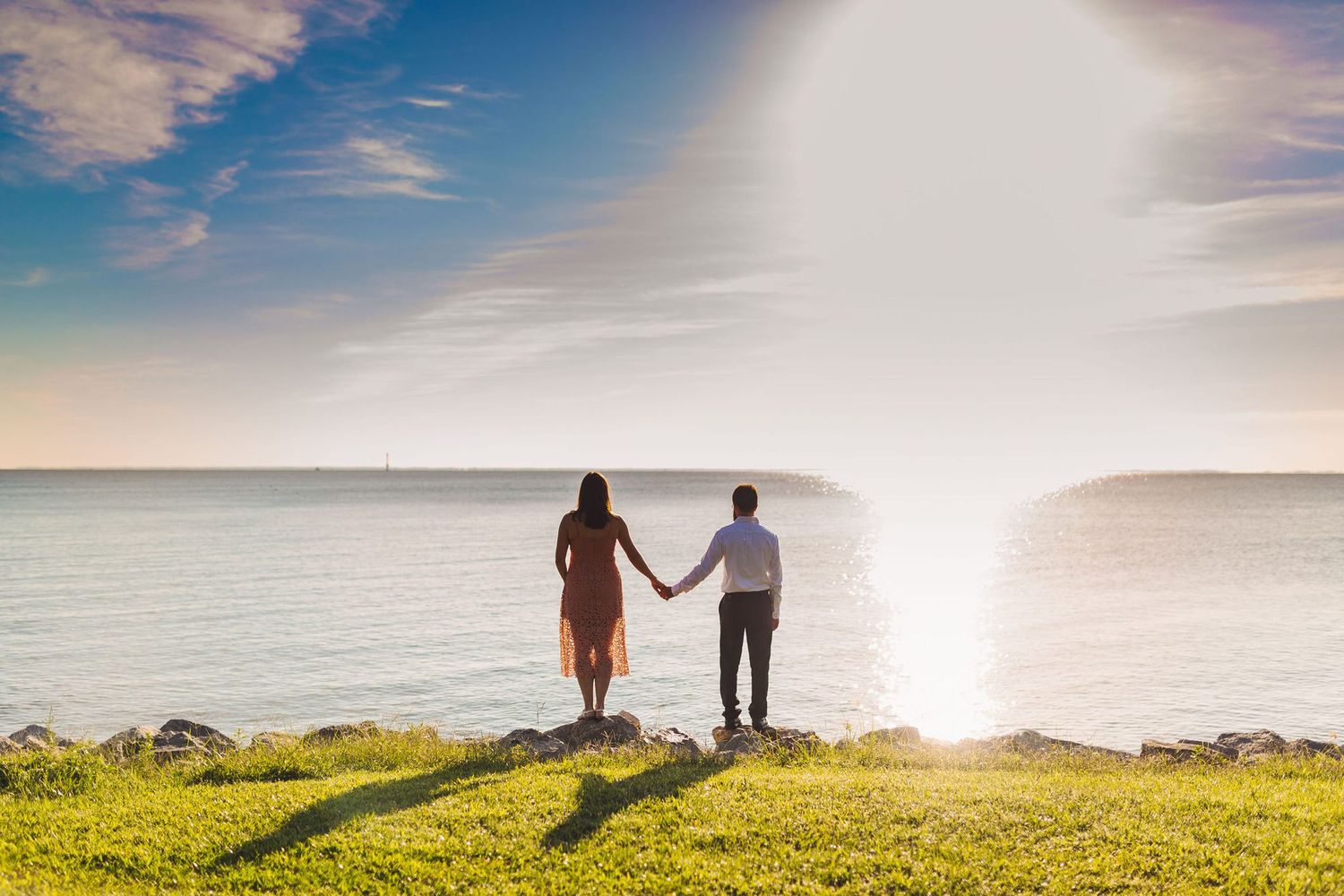 Couple holding hands standing on green cliff overlooking ocean at golden sunset.