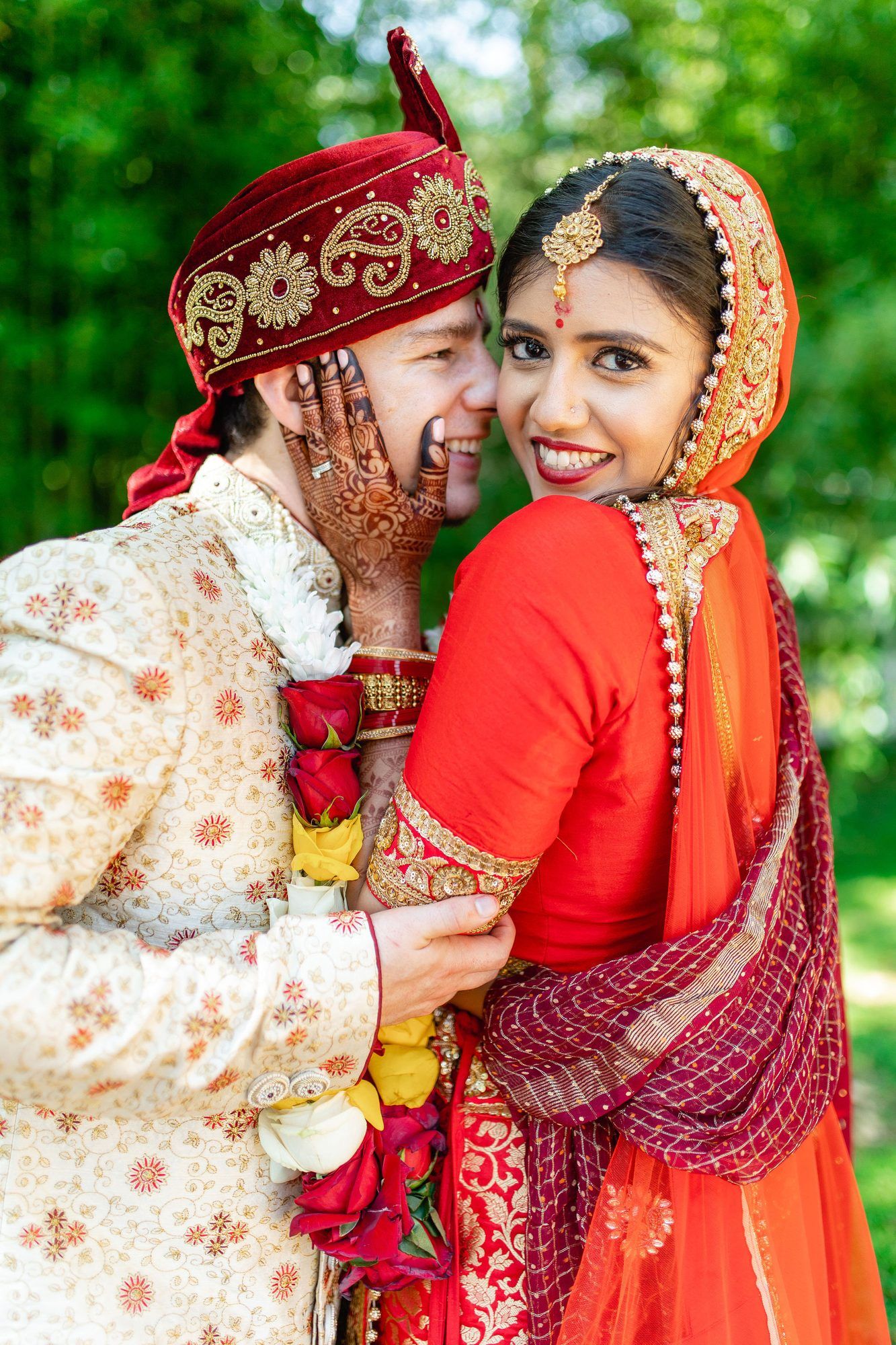 Indian bride and groom in traditional red and gold wedding attire smiling together outdoors.