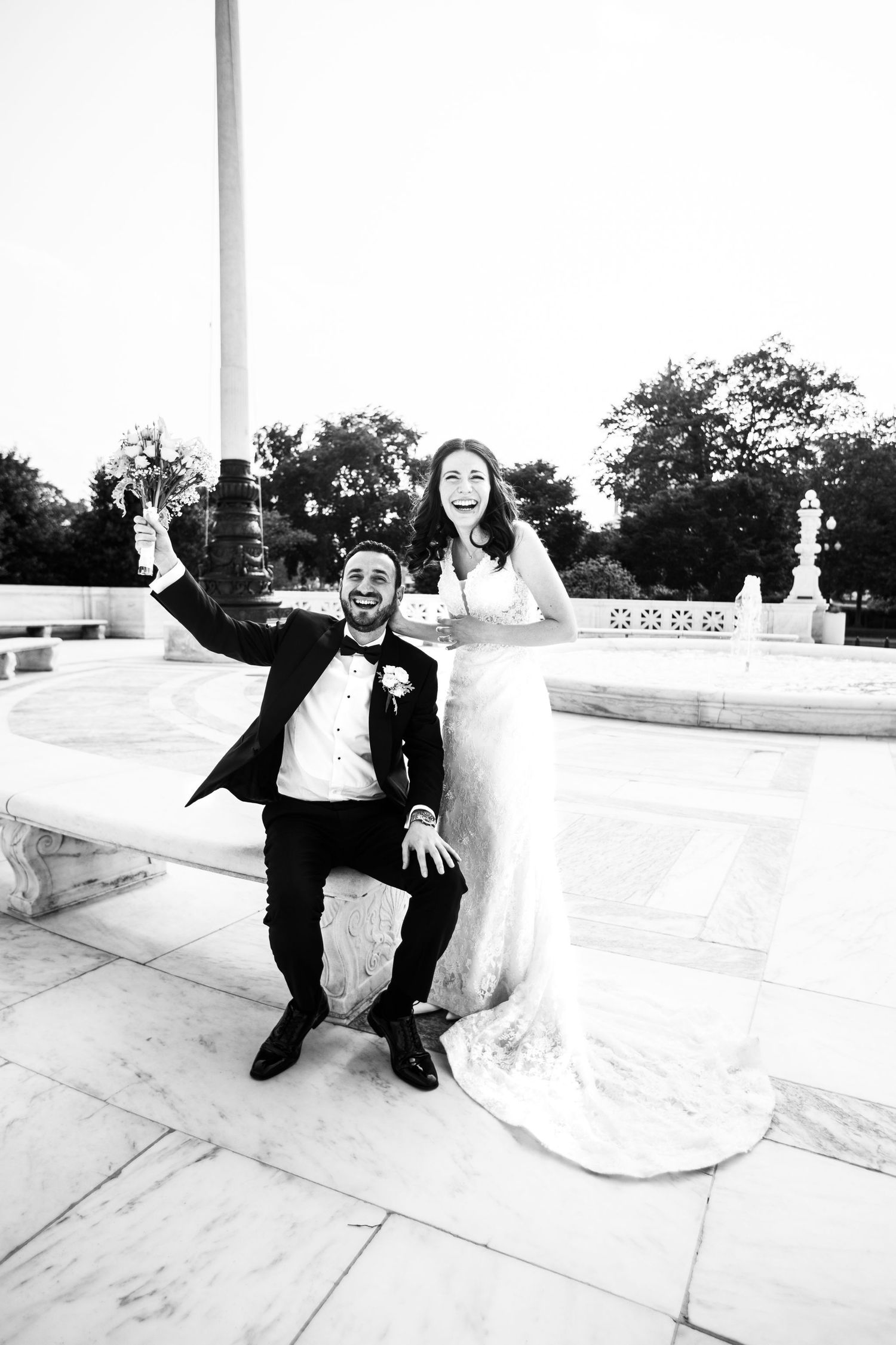 Black and white photo of joyful newlyweds celebrating on marble steps, groom raising arm, bride in wedding gown laughing.