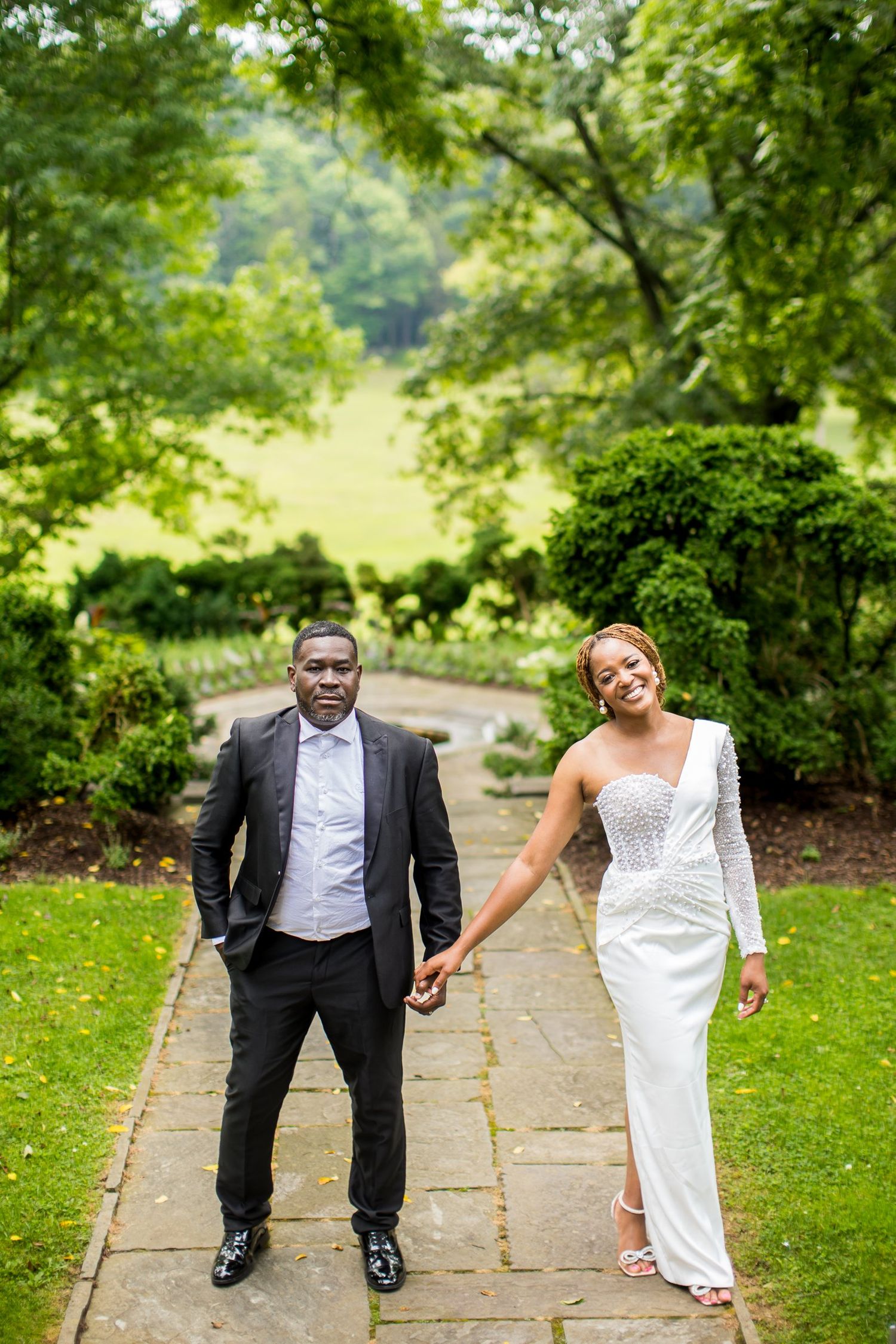 A couple in wedding attire holds hands on a garden path, the bride in a white gown and groom in a dark suit.