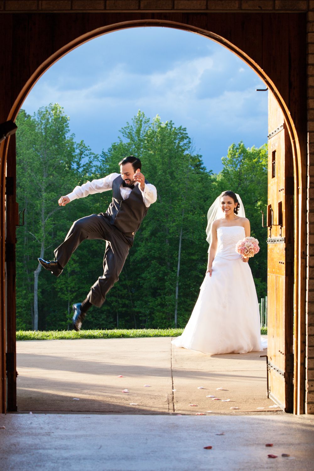 Joyful groom jumping in archway doorway while smiling bride in white gown holds bouquet outdoors at wedding venue.