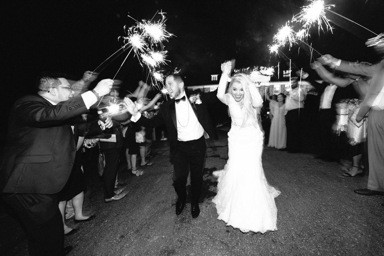 Newlyweds walk through sparkler exit tunnel surrounded by cheering guests at night wedding reception.