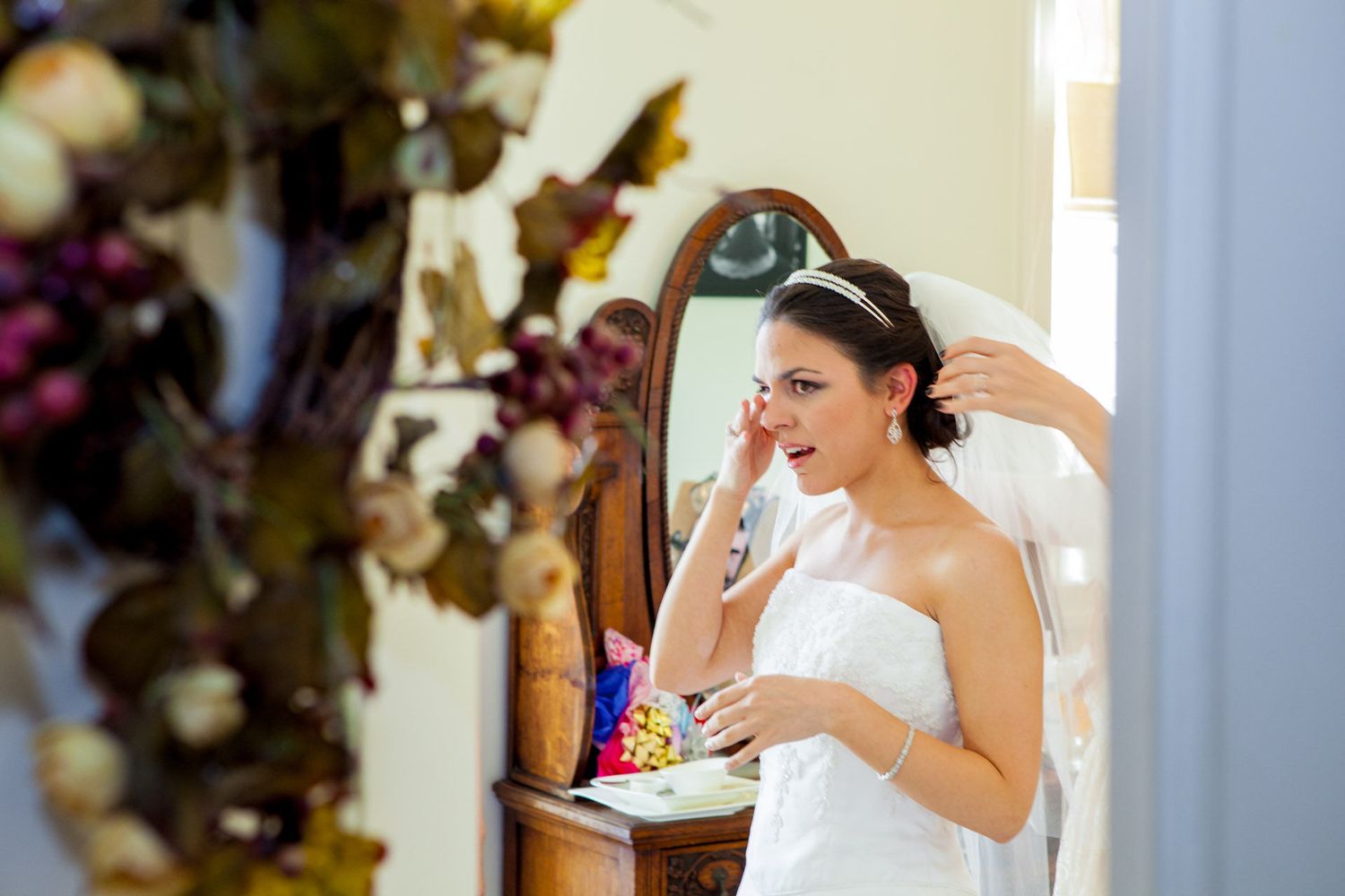 Bride in white dress applying makeup at vintage vanity mirror, preparing for wedding day.