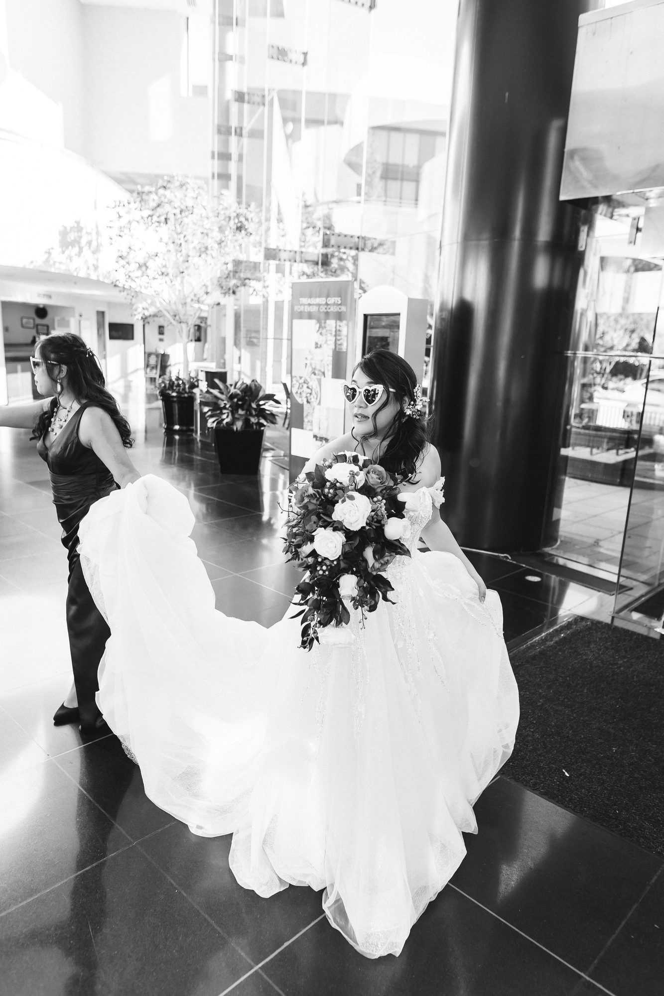 Black and white photo of a bride in a flowing gown and sunglasses holding a bouquet in an elegant hotel lobby.