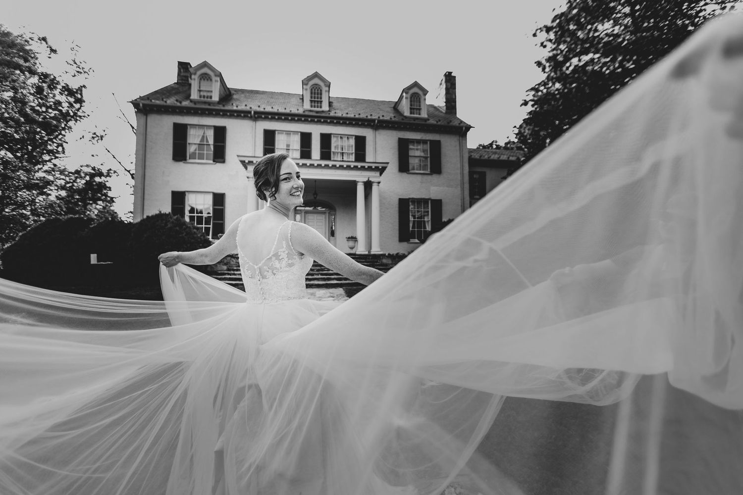 Black and white bridal portrait with flowing veil in front of elegant historic manor house.