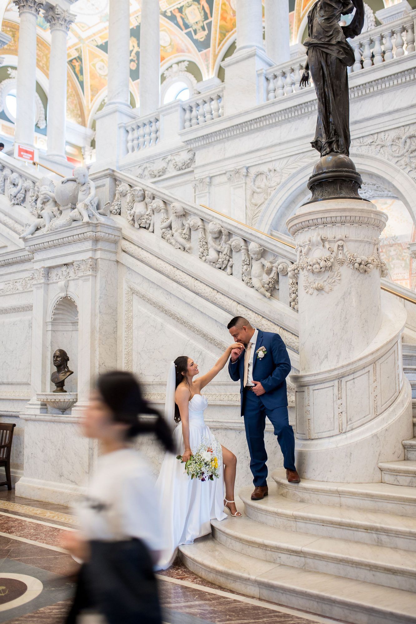 Bride and groom pose on ornate marble staircase in grand hall, with bronze statue and classical architecture.