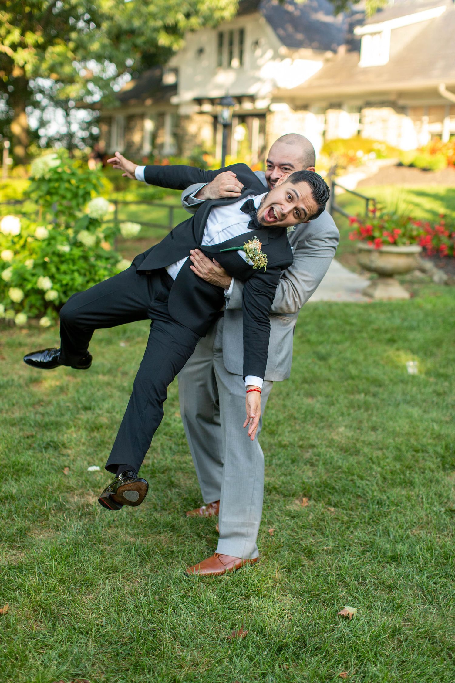 Two grooms in suits laugh and play outside at their wedding reception on a green lawn.