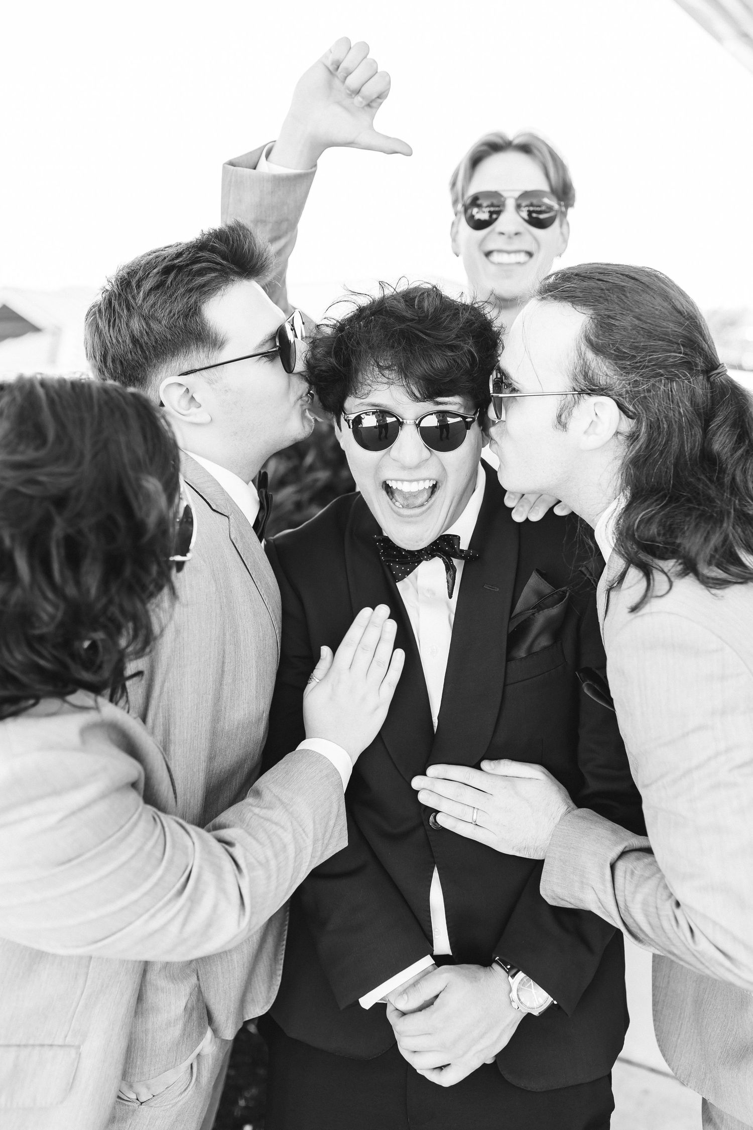 Black and white photo of a groom in a tuxedo laughing as groomsmen in suits surround and celebrate him.