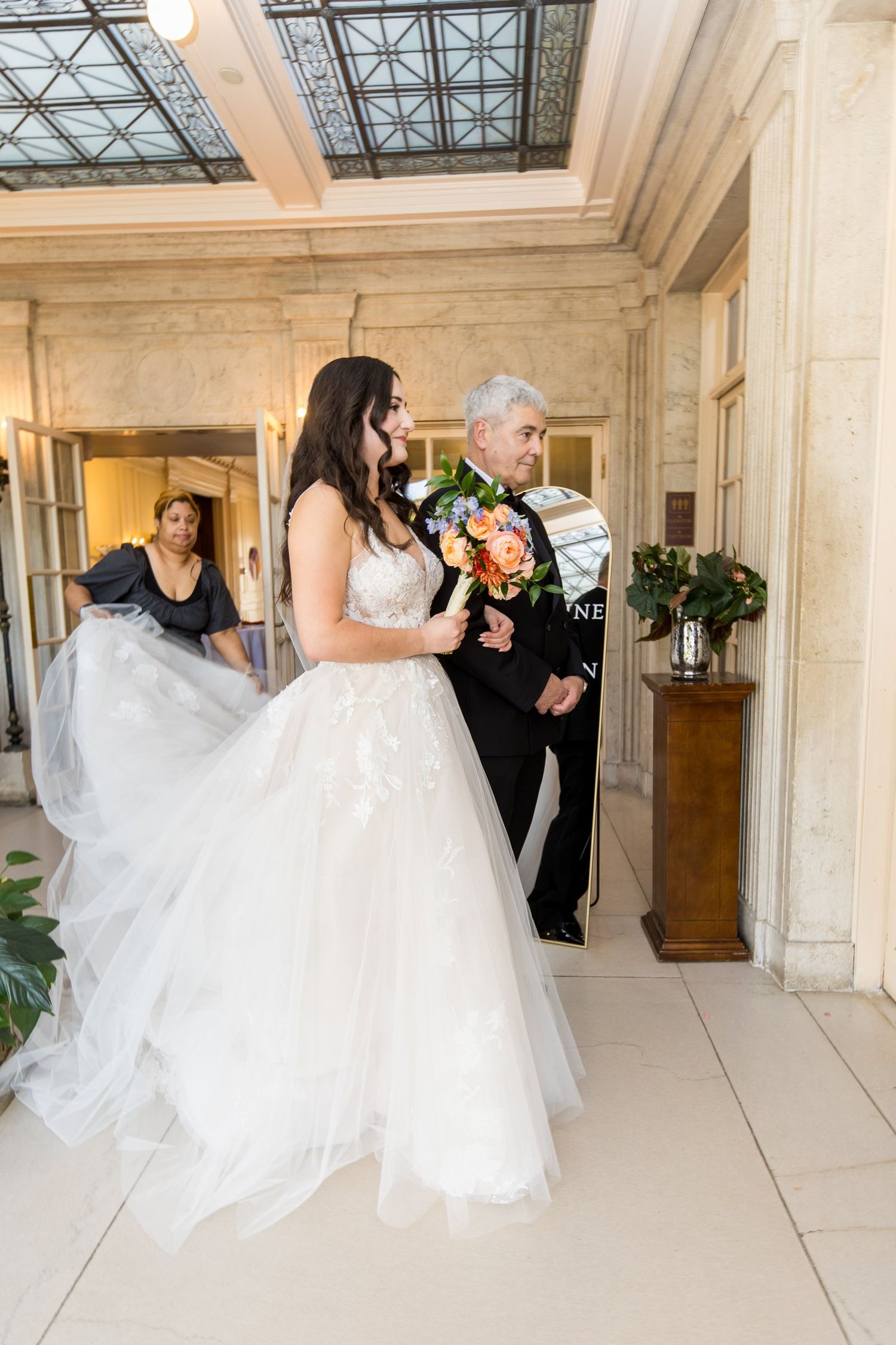Bride in white tulle gown holding colorful bouquet walks with escort through elegant marble venue doorway.